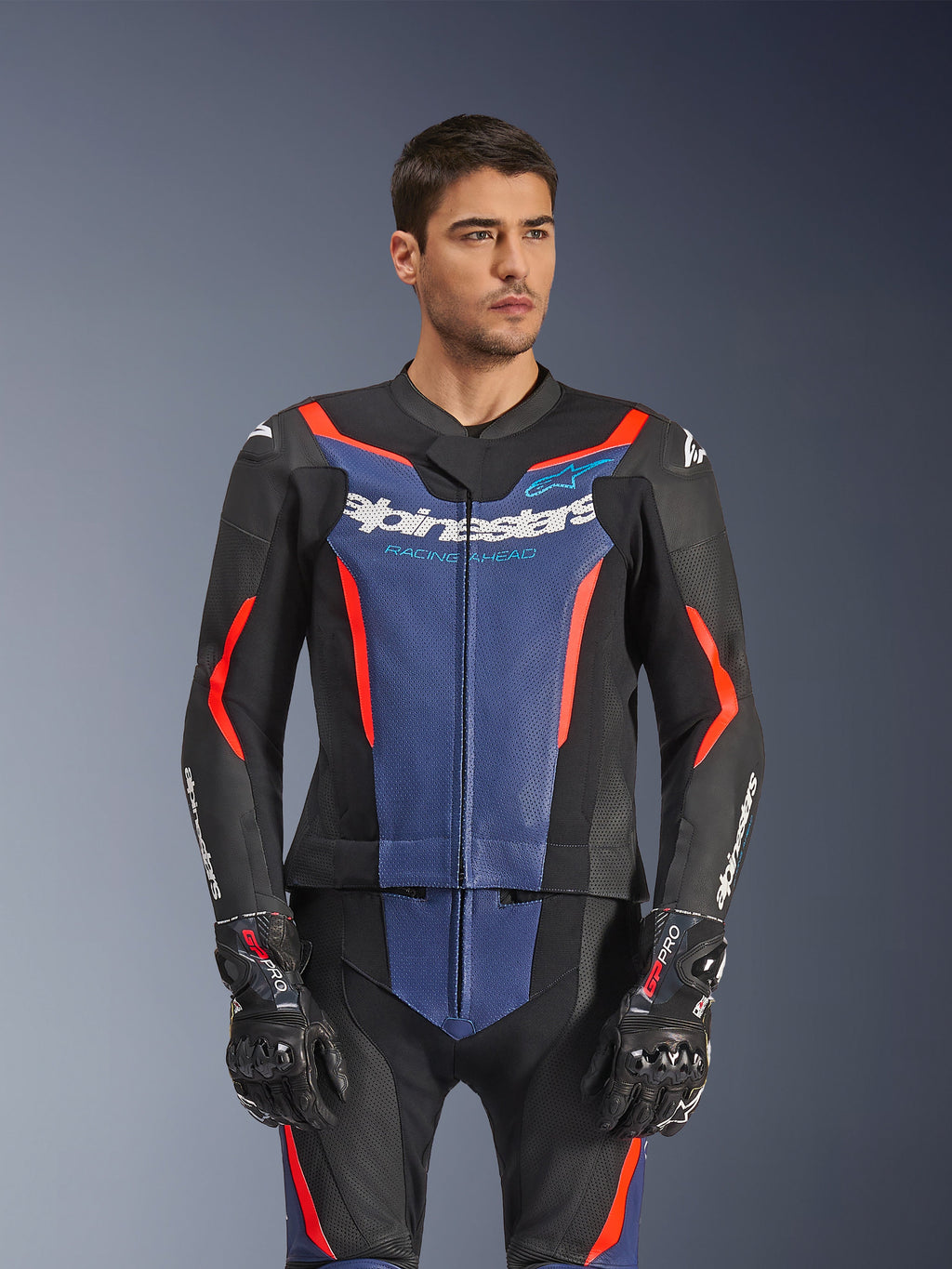 Male model wearing Alpinestars GP Force V2 Leather Suit 2-Piece, Black Blue Red Fluo, featuring bold blue chest panel and red accents, worn with GP Pro gloves, professional leather motorcycle racing gear with perforated texture