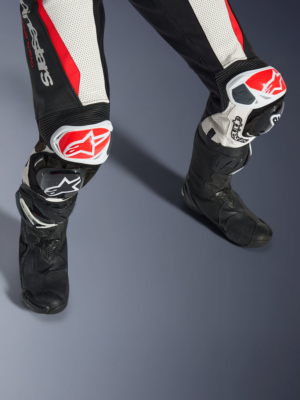 Alpinestars Woman Stella GP-R7 2-Piece Leather Suit, Black White and Red Fluo, close-up of legs and knees featuring red and white GP DFS RR sliders, perforated leather construction with accordion stretch panels, worn with black performance racing boots