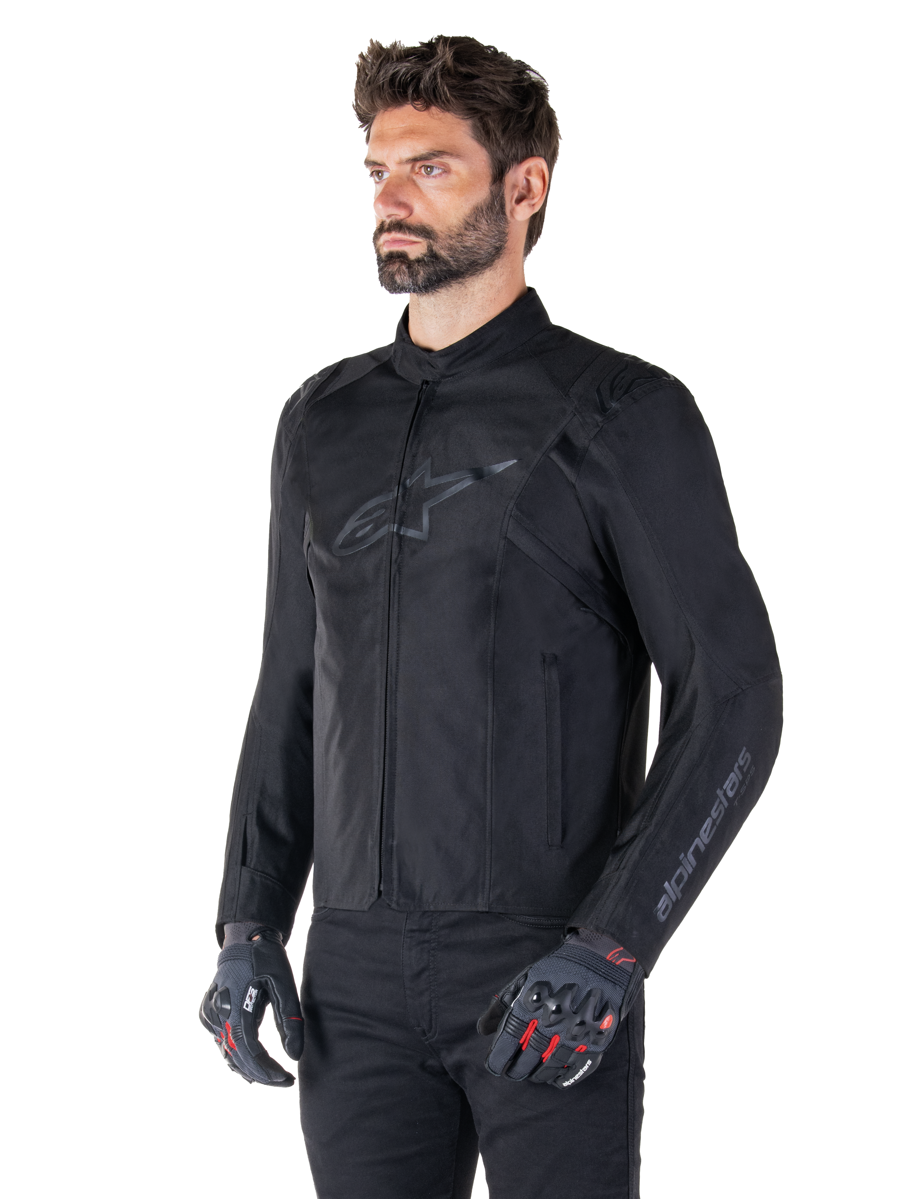 Model wearing Alpinestars T-SPS V2 Waterproof Jacket, Black, paired with black pants and grey motorcycle gloves with red accents, featuring a subtle black chest logo and sleeve branding, textile sport-cut jacket for road riding.