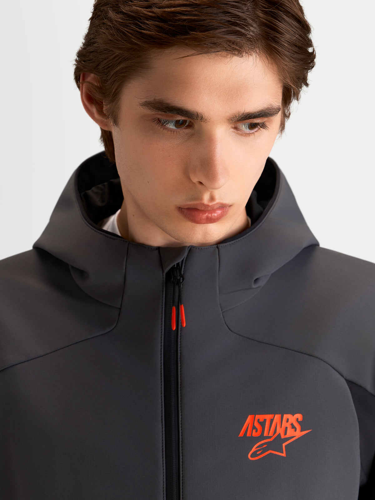 Alpinestars Aeroshell Waterproof Jacket, Men's Motorcycle Sport Softshell Jacket in Black and Dark Gray, featuring an open-slot hood and orange ASTARS logo detail, close-up on model