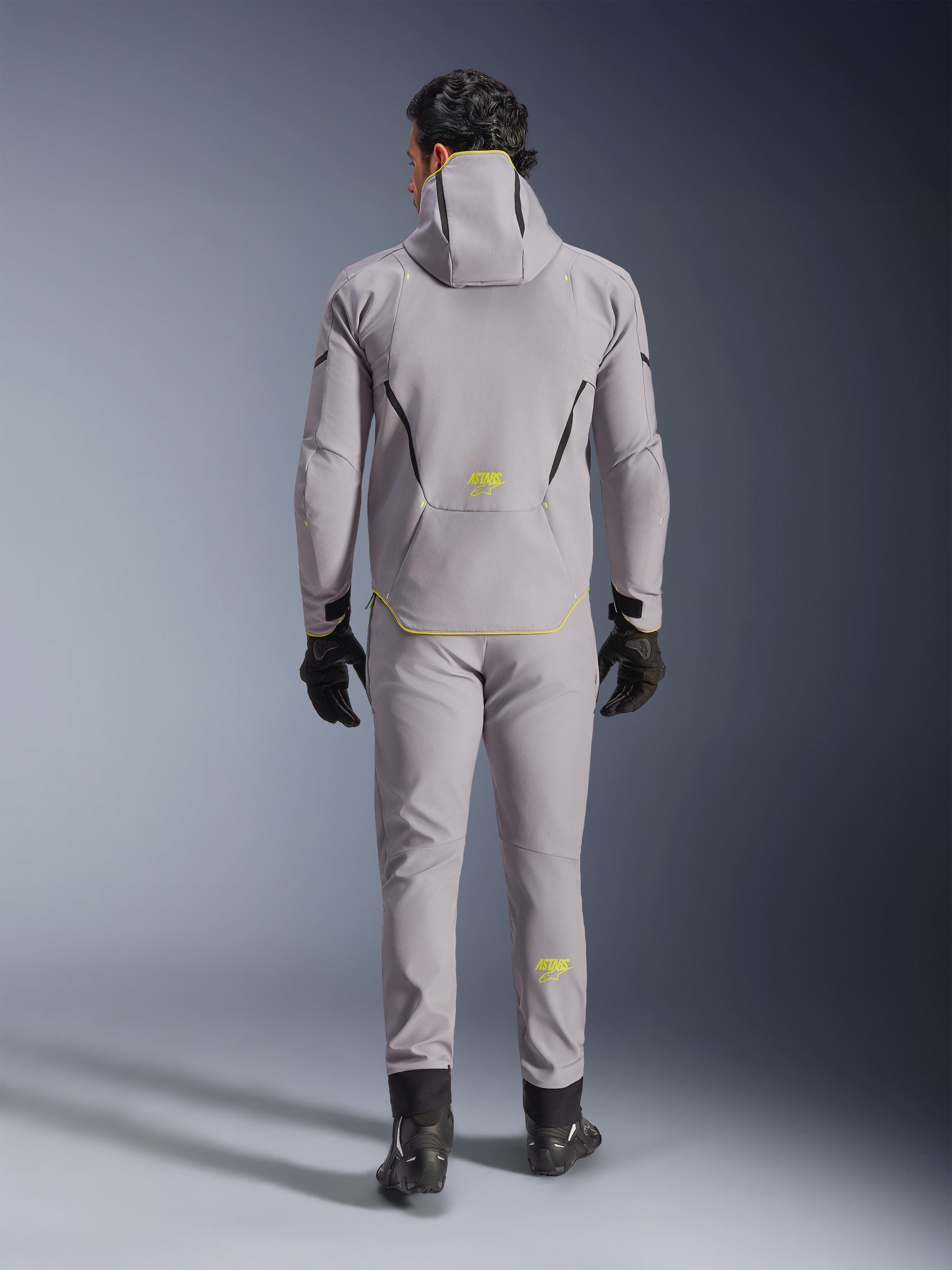 Alpinestars Aeroshell Waterproof Jacket in Mid Gray Yellow Fluo, full back view, worn with matching gray pants, black gloves, and black riding shoes. Features a hooded softshell design with yellow fluo 'ASTARS' branding and accents, engineered for modern motorcycle road performance.