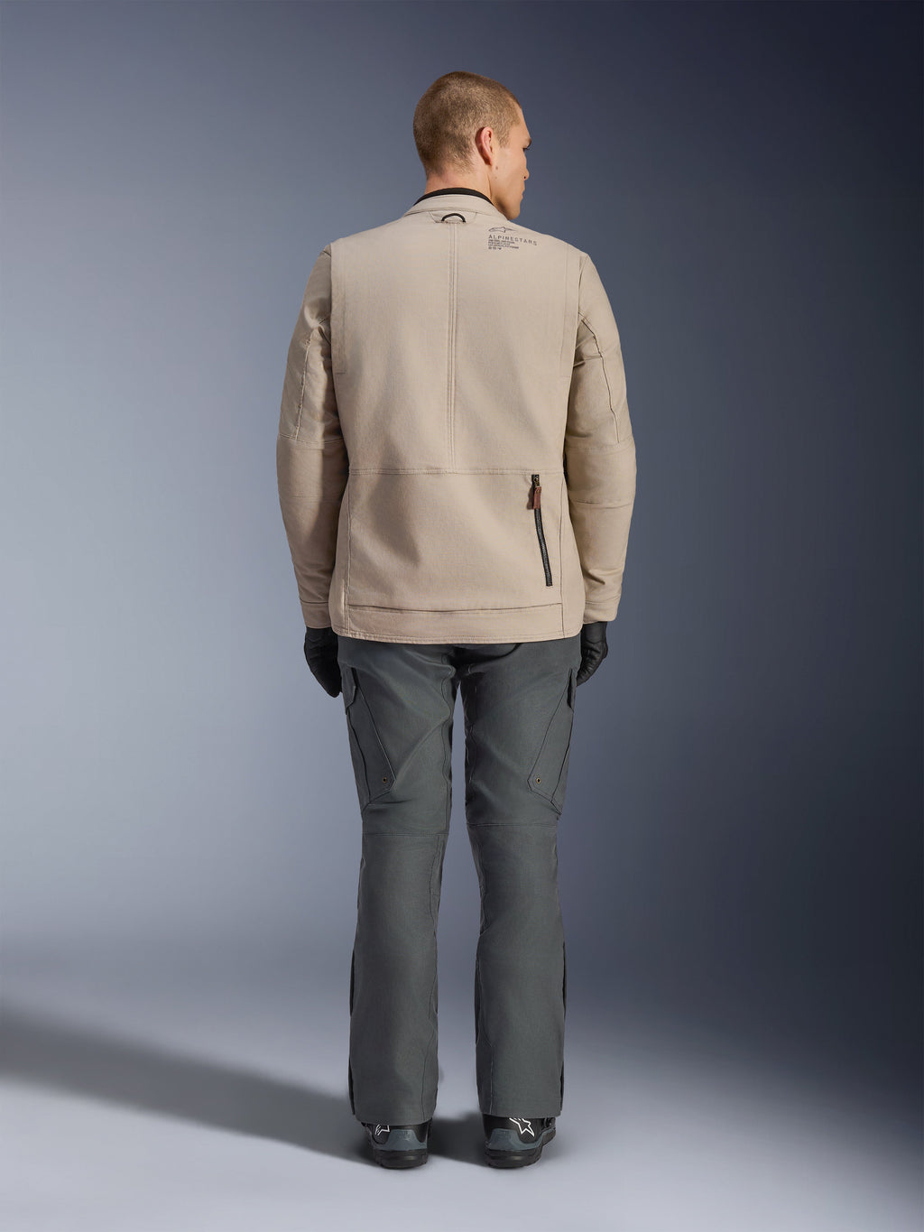 Alpinestars Borrego Drystar® Jacket, back view of male model wearing the Warm Gray textile motorcycle jacket, paired with dark gray technical pants, black gloves, and boots, featuring the rugged waxed-canvas design and rear expansion panels.