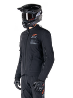 Alpinestars AMT-8 Stretch Drystar® XF Jacket, Black, worn by a rider with matching adventure pants and gloves, featuring a black motocross helmet and goggles, zippered ventilation ports on the chest and arms, Alpinestars Motion Technology branding with an orange logo, premium adventure-touring gear