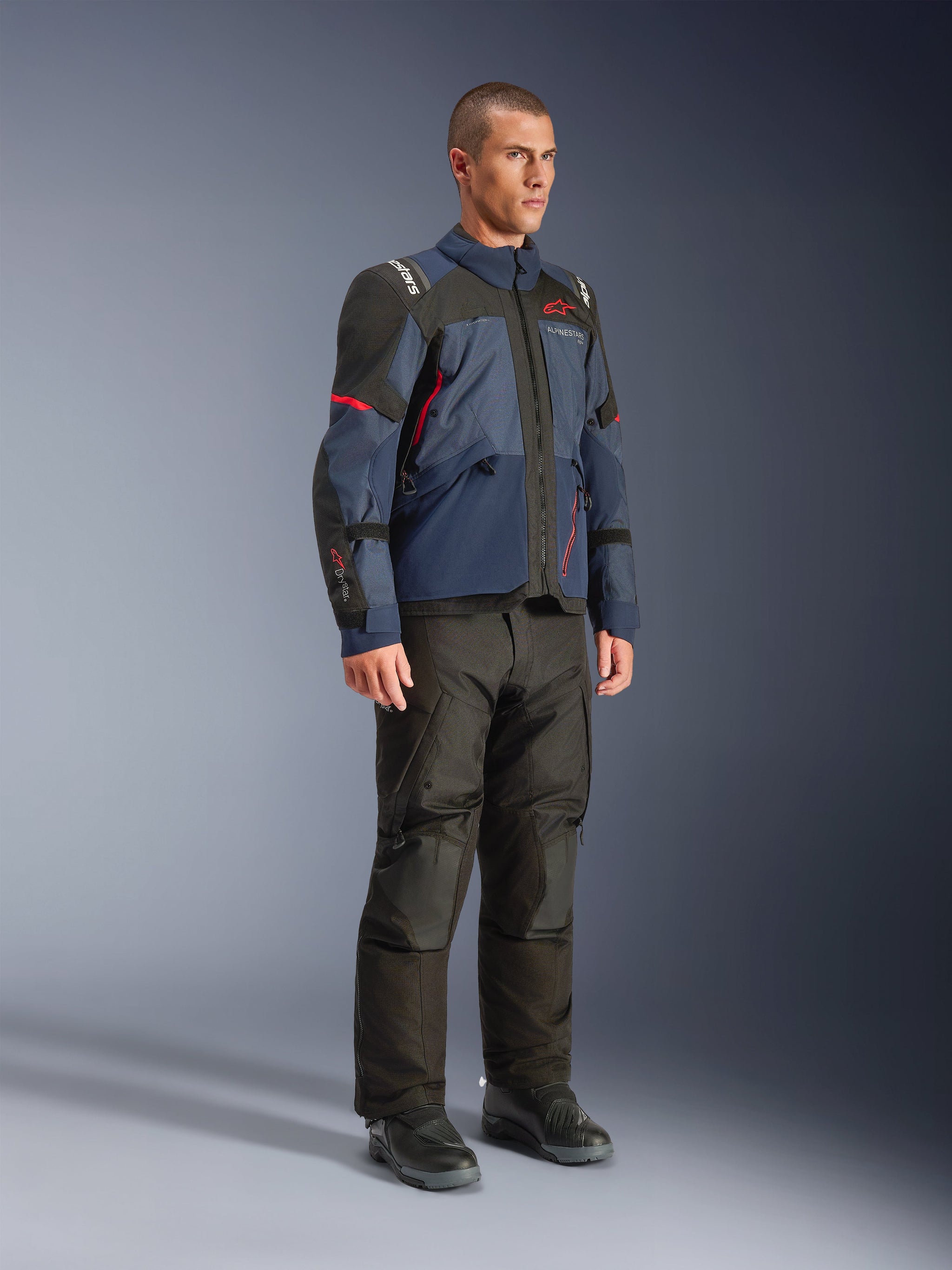 Male model wearing Alpinestars Andes V4 Drystar® Jacket in Night Navy and Mood Indigo with black panels and red accents, paired with black motorcycle pants and boots, technical adventure touring gear in a studio setting.