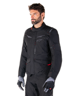 Alpinestars ST-1 Waterproof Jacket, Black with red accents, worn by a male rider with grey and black gloves, featuring a minimalist adventure touring design, cargo pockets, and subtle Alpinestars branding.