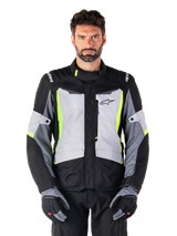 Alpinestars ST-1 Waterproof Jacket, adventure touring motorcycle jacket, Ice Gray Black and Yellow Fluo, worn by a man with matching gloves and black pants, featuring neon yellow accents on the chest and collar, Alpinestars logo on the chest