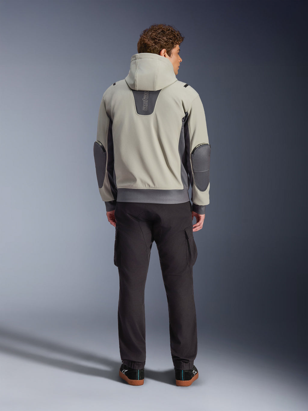 Alpinestars Moblast Waterproof Jacket, Iron Gray Dark Sage, rear view of a male rider wearing the hooded motorcycle jacket, light gray and dark sage colorway, vertical 'astars' logo on the upper back, dark gray elbow patches, styled with dark cargo pants for an urban contemporary look.
