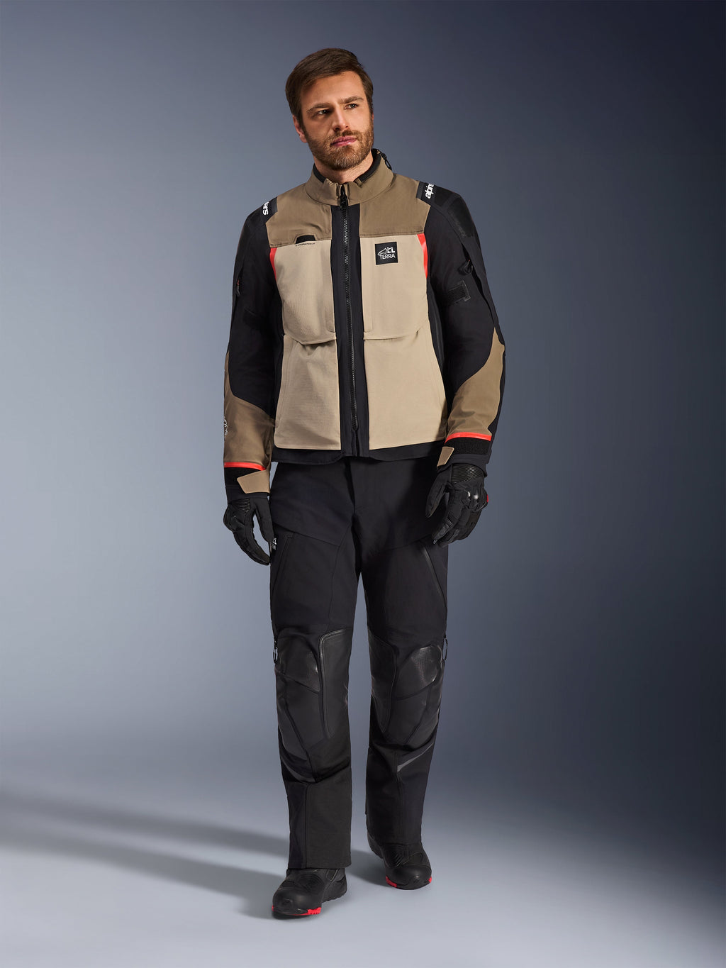 Male model wearing Alpinestars Andes Pro Drystar® XF Jacket in Walnut Aluminum, tan and black adventure touring jacket with red accents, styled with black motorcycle pants, gloves, and boots, full-body studio shot for protective riding gear.