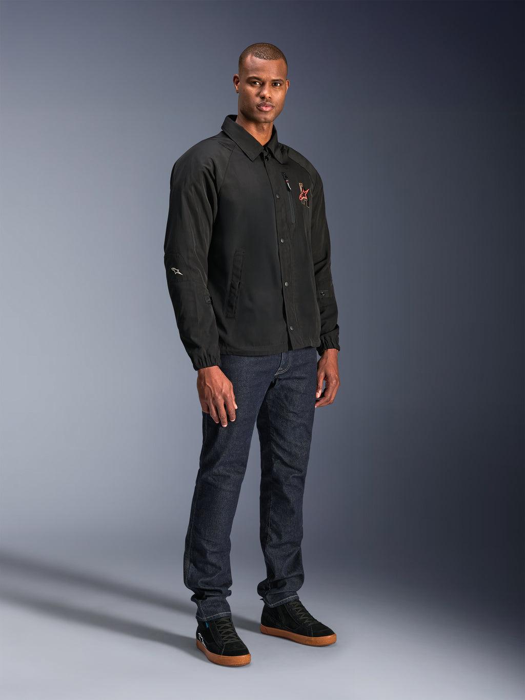Alpinestars Revved WR Jacket in Black, urban coaches-style motorcycle jacket worn by a model with dark blue jeans and black sneakers, featuring a snap-button front and embroidered logo, minimalist and sporty design for road riding.