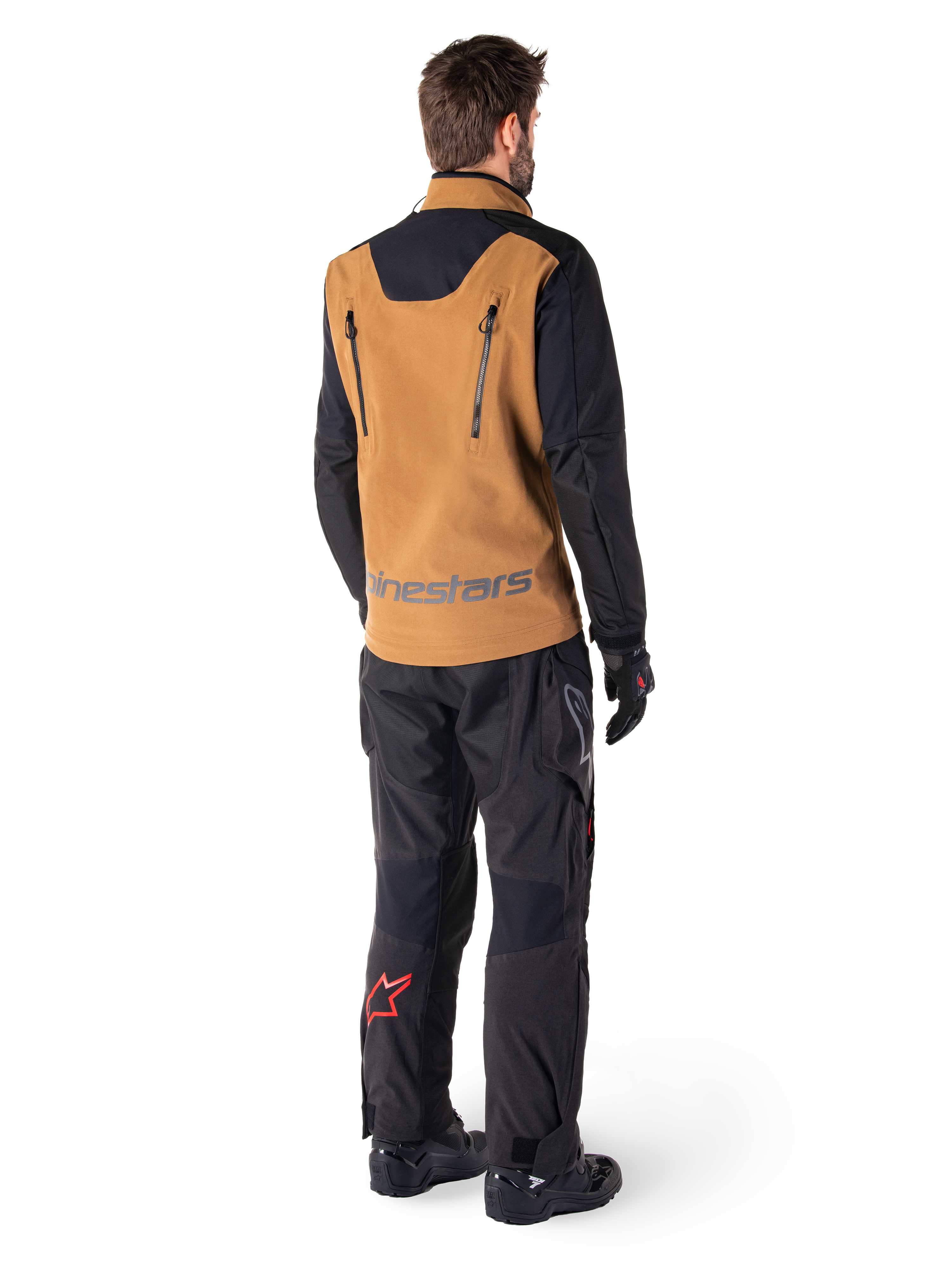 Alpinestars Hyde XT Stretch Drystar®XF Jacket and Hyde XT Drystar® Pants, rear view of a rider wearing tan and black adventure touring gear, jacket featuring vertical back zippers and Alpinestars branding, black pants with red star logo on the calf, paired with black gloves and boots.