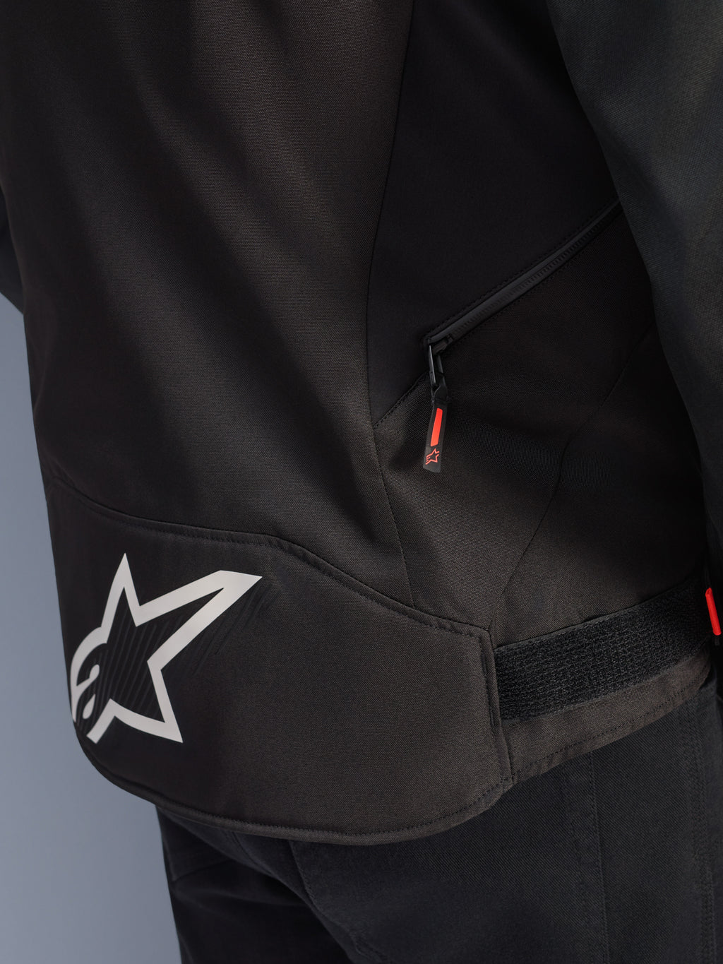 Alpinestars T-GP R V3 Drystar® Jacket, Black and White, close-up view of the lower back section featuring a large white Alpinestars star logo, a side zippered pocket with a red pull tab, and a velcro waist adjustment strap, durable textile material.