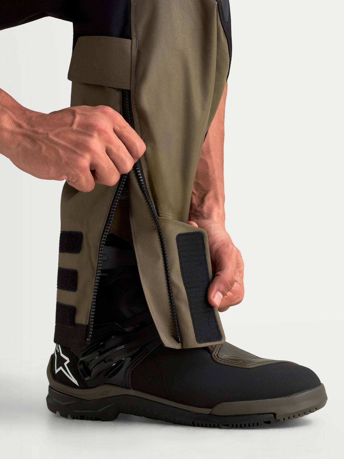 Alpinestars Harok V2 Drystar Pants, men's adventure touring motorcycle pants in aluminum, walnut, and black, close-up detail view of the over-the-boot lower leg construction with a zippered closure and adjustable strap being handled by a rider