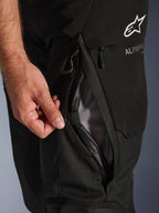 Close-up of Alpinestars Andes V4 Drystar® Pants in Black, featuring a hand opening the direct thigh ventilation port with mesh lining, highlighting the durable textile construction and Alpinestars branding.