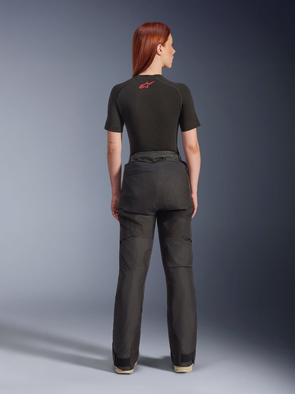 Rear view of a model wearing Alpinestars Stella Andes V4 Drystar® Pants in Black, paired with a black short-sleeve base layer featuring a red Alpinestars logo, adventure-touring motorcycle gear with durable textile construction.