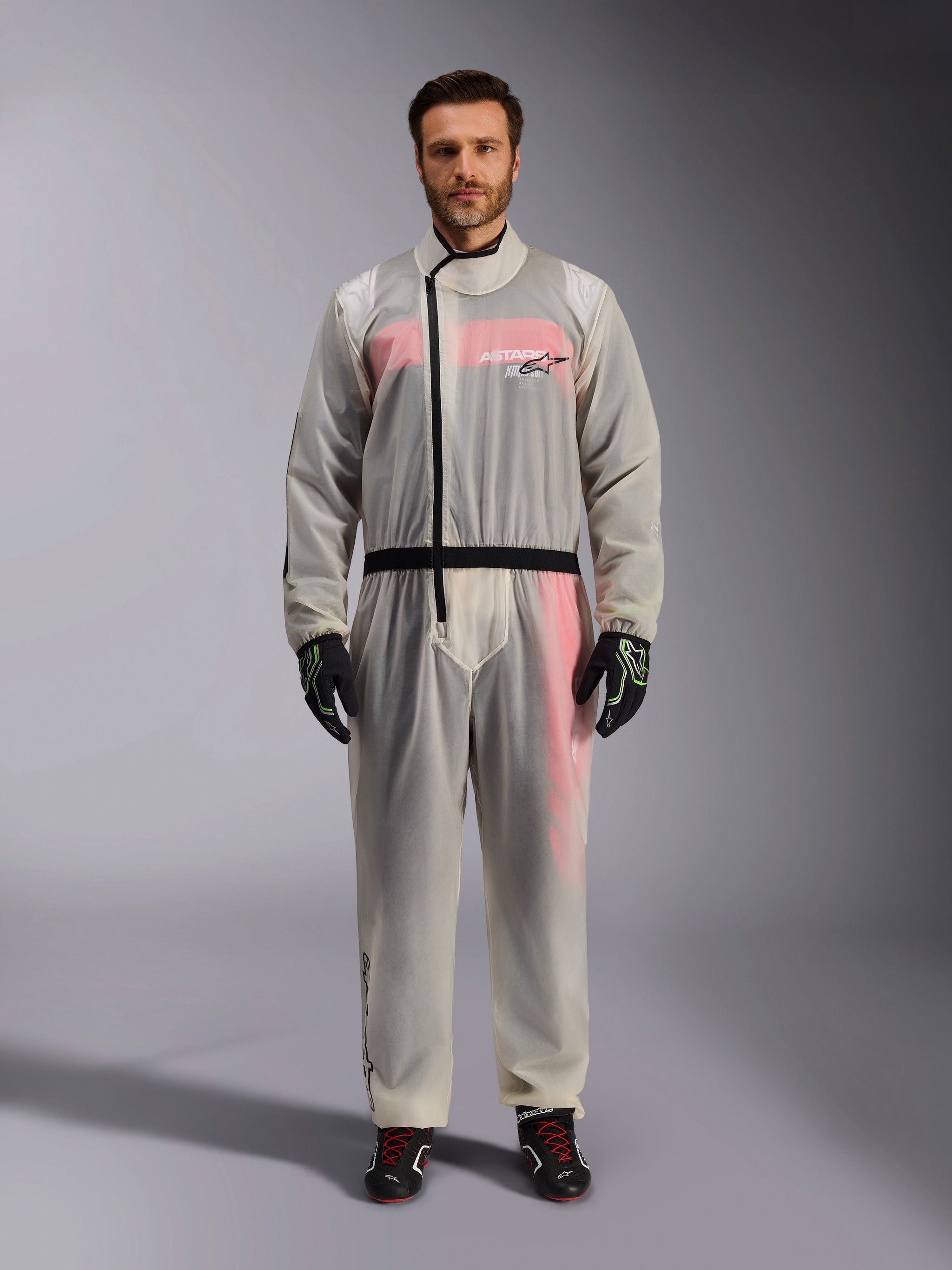Alpinestars Kart Rain Suit, Clear, transparent waterproof oversuit worn by a model, featuring a black asymmetric zipper and chest logo, red and white racing suit visible underneath, paired with black and neon green gloves and black racing boots.