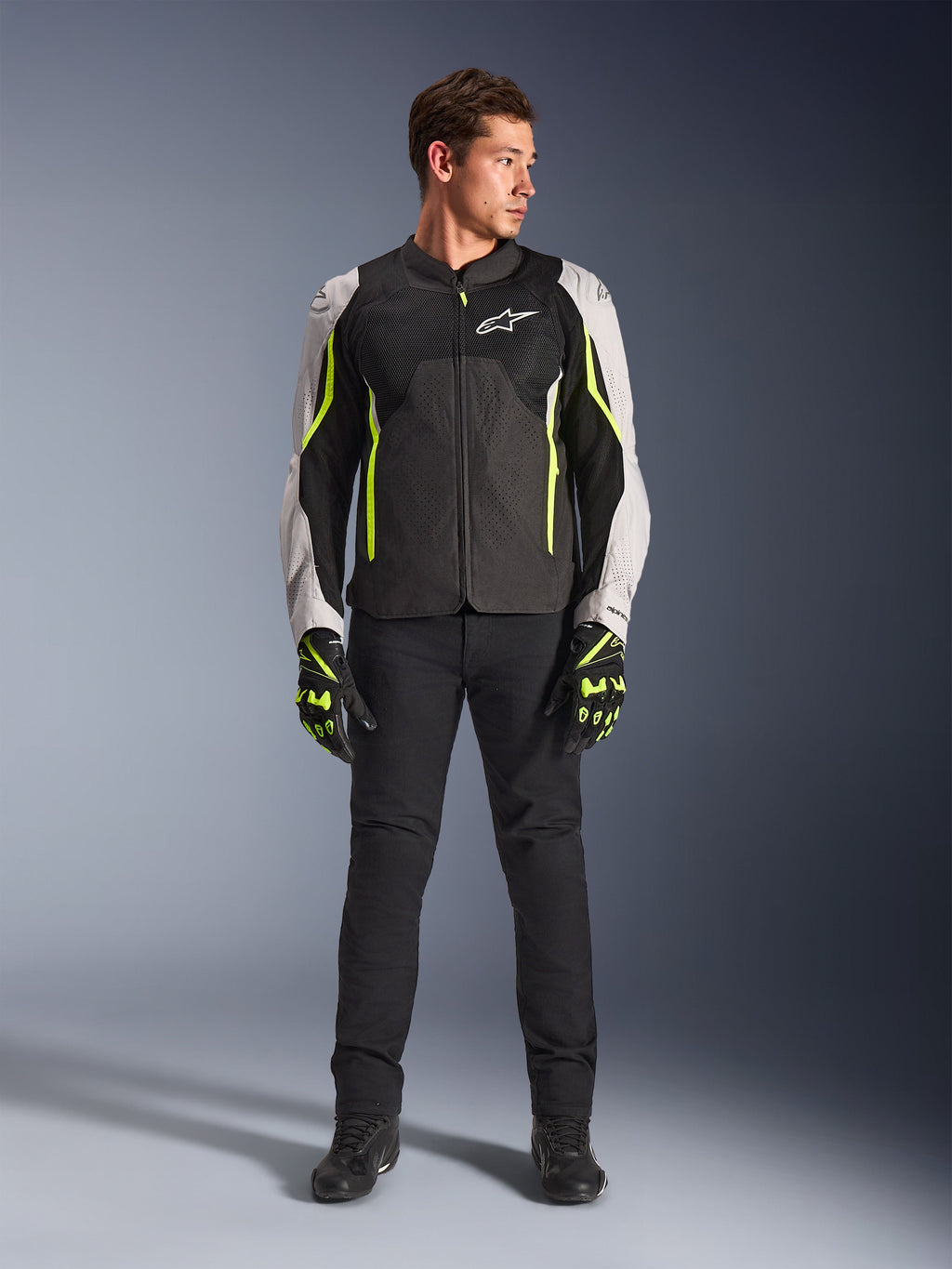 Male model wearing Alpinestars AST V3 Air Jacket in Black Silver Yellow Fluo, featuring mesh ventilation panels and fluorescent accents, paired with black riding pants and matching motorcycle gloves, sport-riding performance gear.