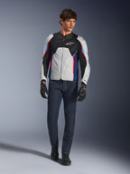 Male model wearing the Alpinestars AST V3 Air Jacket in Silver Deep Blue Bright Red, featuring mesh ventilation and perforated panels, paired with dark denim riding jeans and black gloves, sport-optimized motorcycle gear for warm weather.