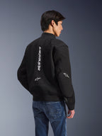 Alpinestars Flight Air Jacket, Black motorcycle jacket, back view showing large vertical white logo and mesh ventilation panels, bomber-style with ribbed hem, worn with blue jeans, urban contemporary riding gear