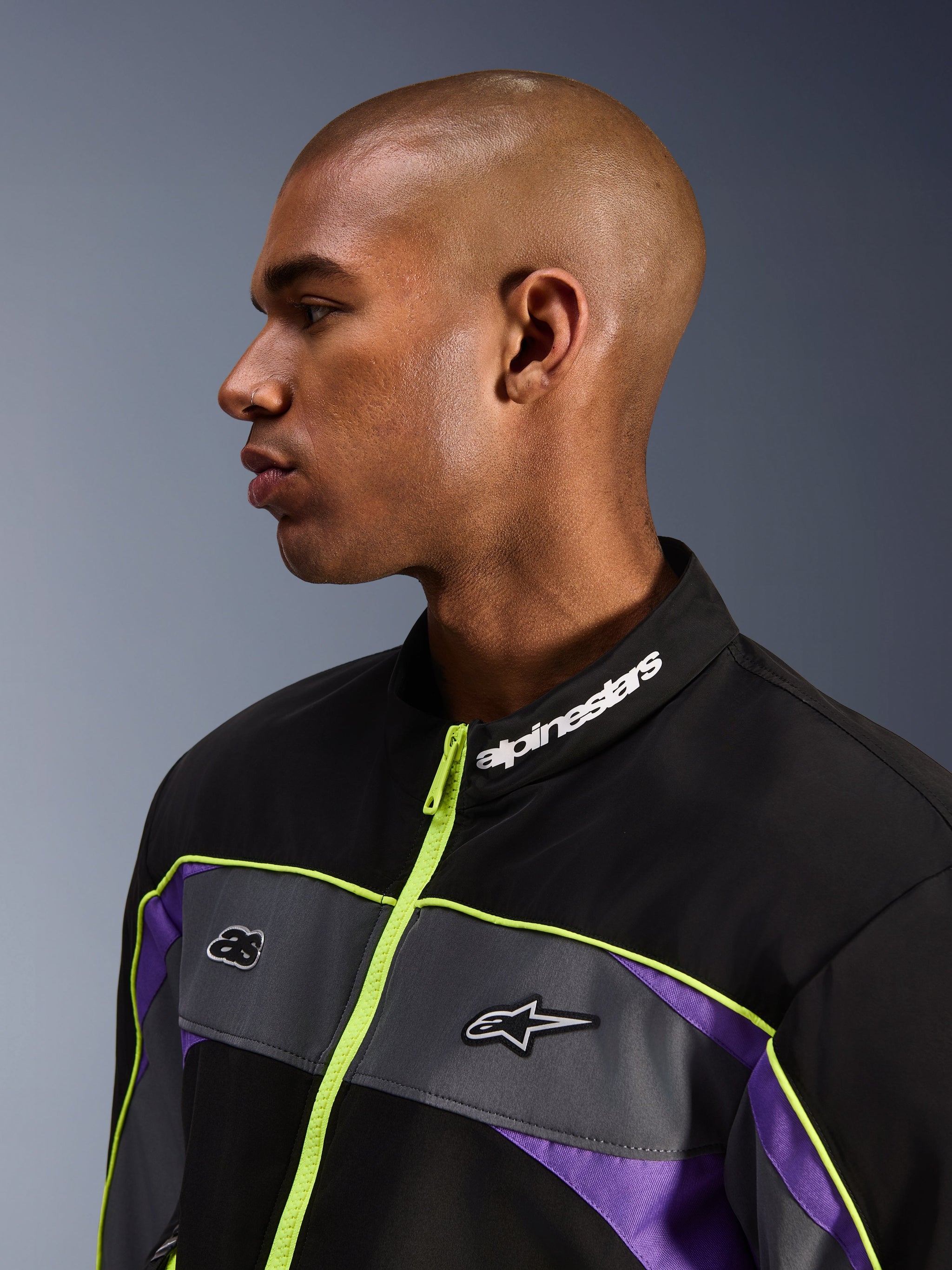 Alpinestars Unite Jacket, Black Asphalt Violet, male model profile view focusing on the high collar with white Alpinestars branding, neon yellow zipper, grey chest panels with logos, purple accents, minimalist urban motorcycle streetwear styling