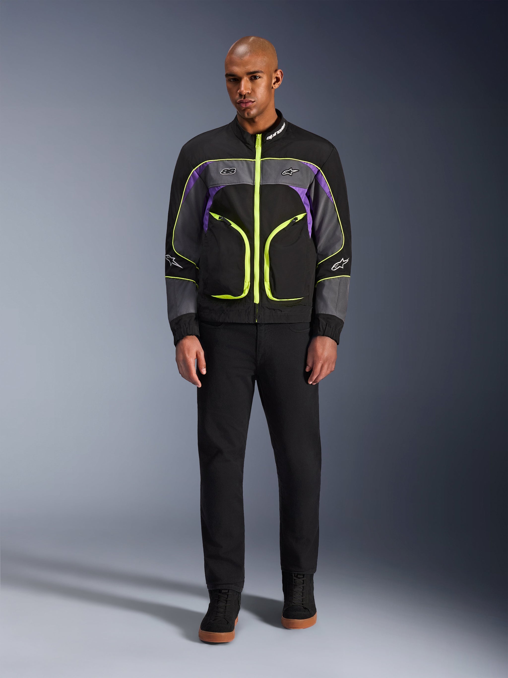 Male model wearing the Alpinestars Unite Jacket in Black Asphalt Violet, featuring a black base with grey and purple panels and vibrant neon yellow zippers and trim, paired with black pants and shoes, minimalist urban motorcycle jacket.