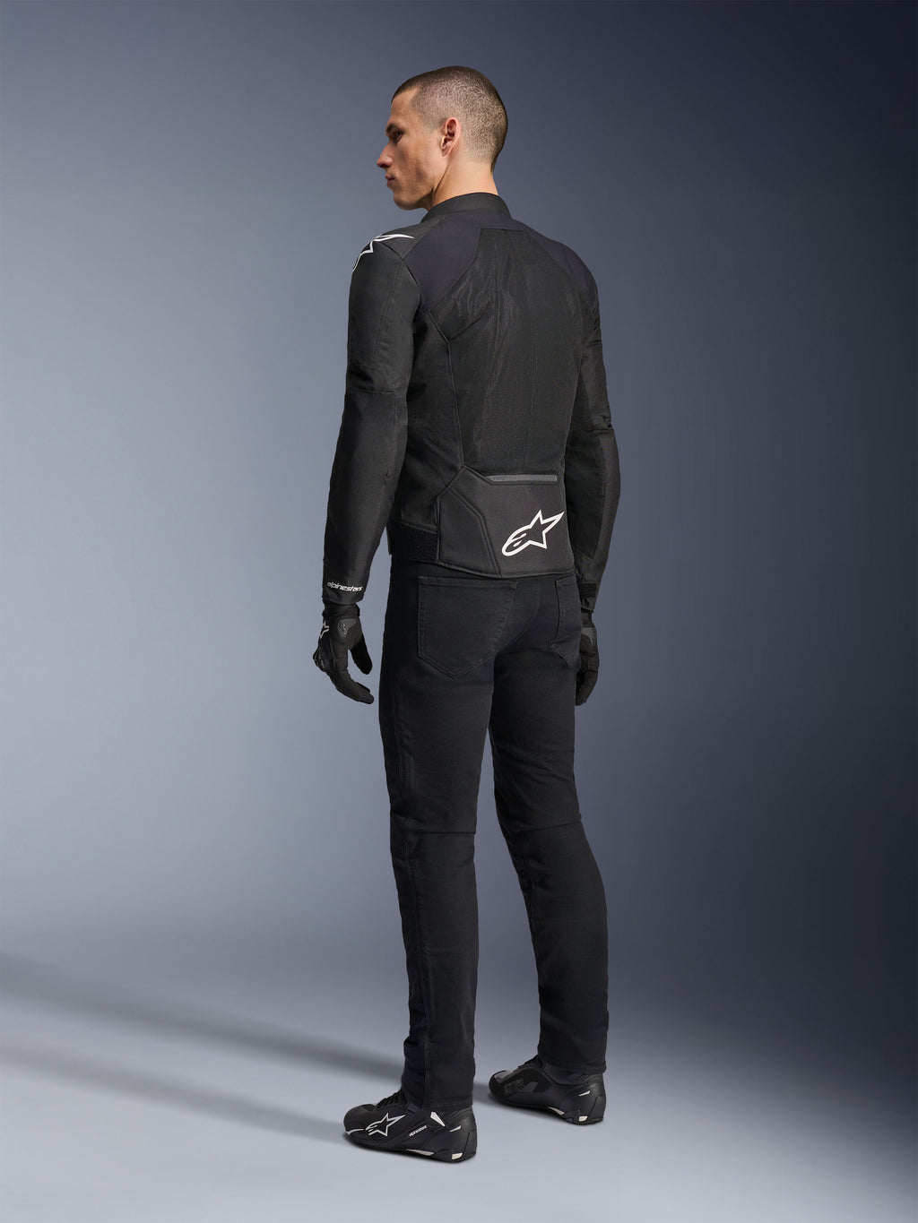 Alpinestars Viper Air V4 Jacket, Black textile motorcycle jacket, rear view on a male model, featuring large tech mesh panels for airflow and a white star logo on the lower back, worn with matching black riding pants and gloves