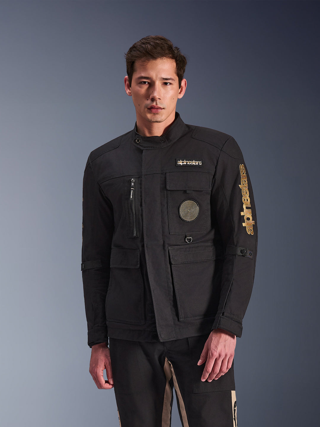 Alpinestars Dice Jacket, Black Motorcycle Jacket, worn by model, featuring gold Alpinestars logos, multiple functional pockets, circular spiral patch, and urban field jacket styling