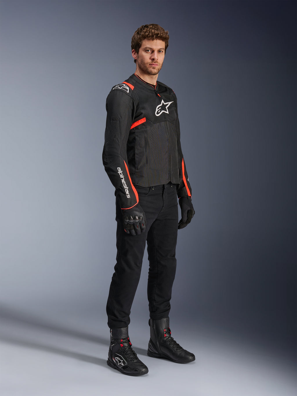 Male model wearing Alpinestars T-Sps Air V2 Jacket, Black with Red Fluo accents, paired with black motorcycle pants, black gloves, and black boots, highlighting a sporty design with mesh panels and white Alpinestars logos.