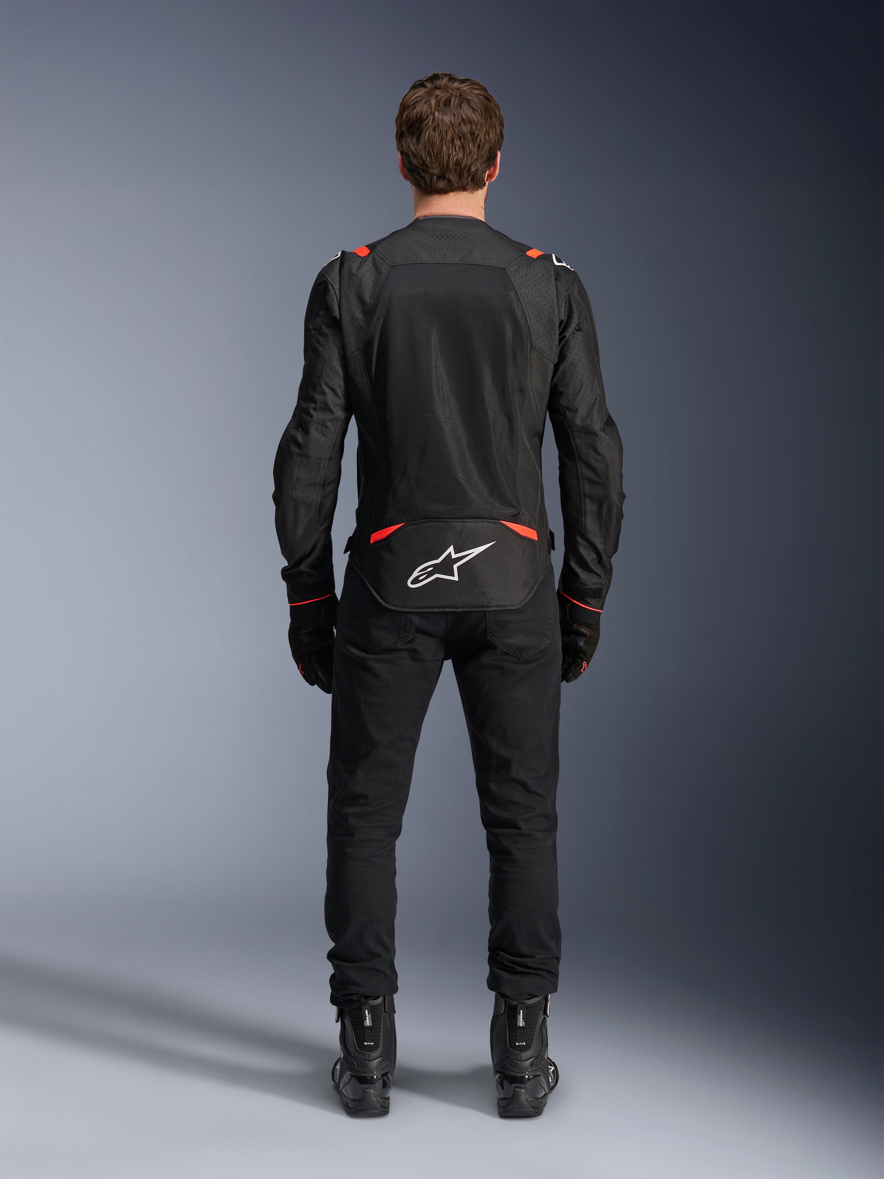 Alpinestars T-SPS Air v2 Jacket in Black and Red Fluo, rear view showing large mesh ventilation panels and white Alpinestars logo on the lower back, worn with black riding pants and boots.