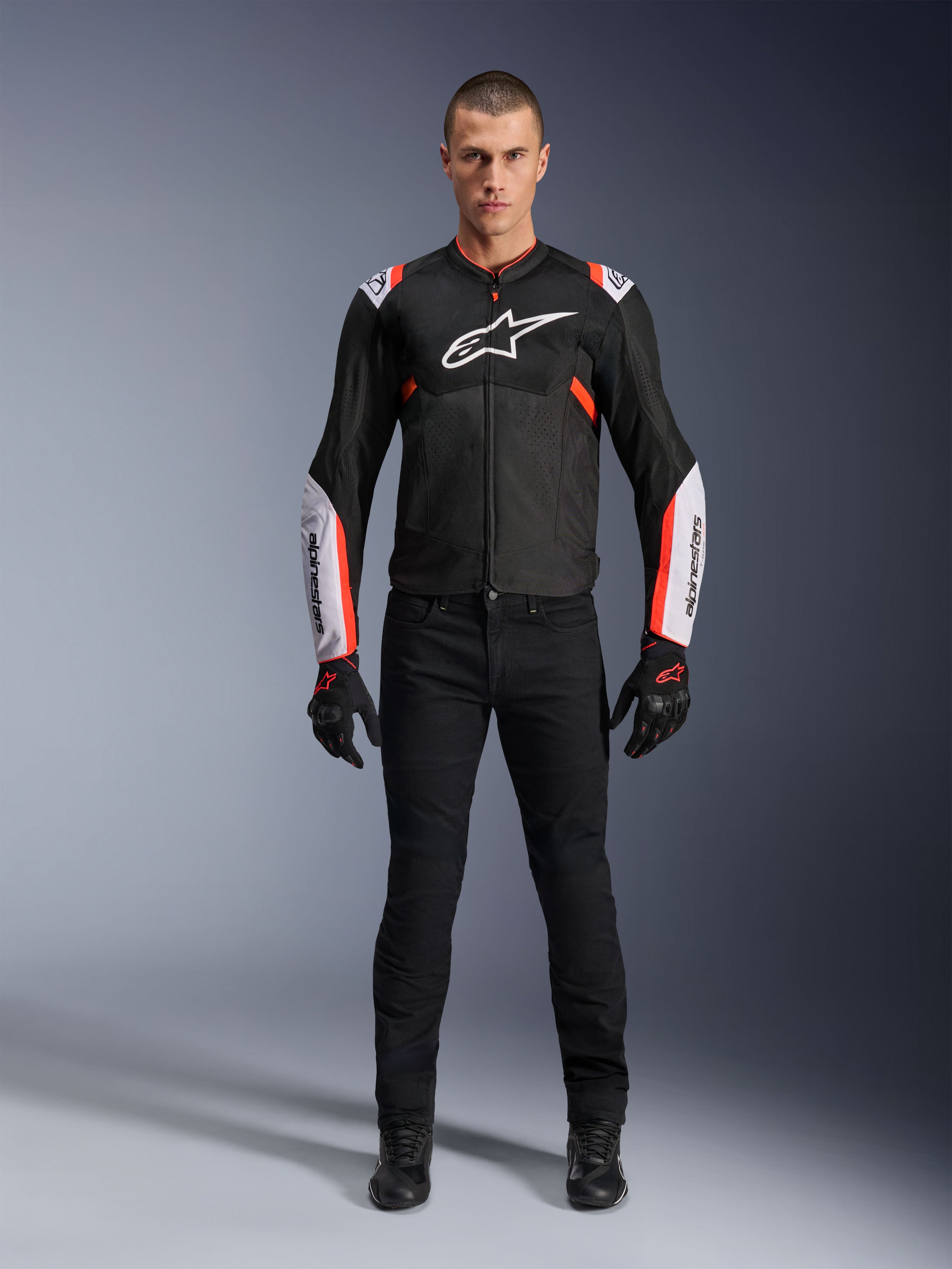 Male model wearing Alpinestars T-Sps Air V2 Jacket in Black White Red Fluo, featuring large tech mesh panels and bold white branding on the chest and sleeves, paired with black technical riding pants and gloves, urban sport motorcycle gear