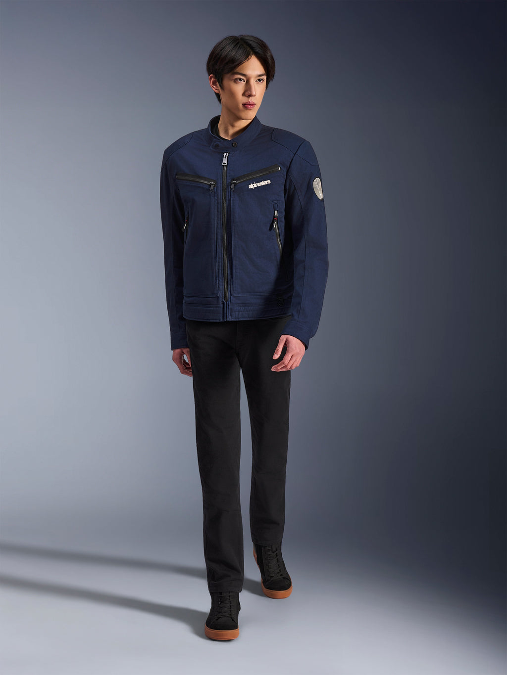 Male model wearing Alpinestars Bruiser Jacket in Dark Blue, urban textile motorcycle jacket, paired with black pants and black sneakers, workwear-inspired aesthetic with zippered pockets and circular sleeve patch, contemporary streetwear for motorcycle riding.
