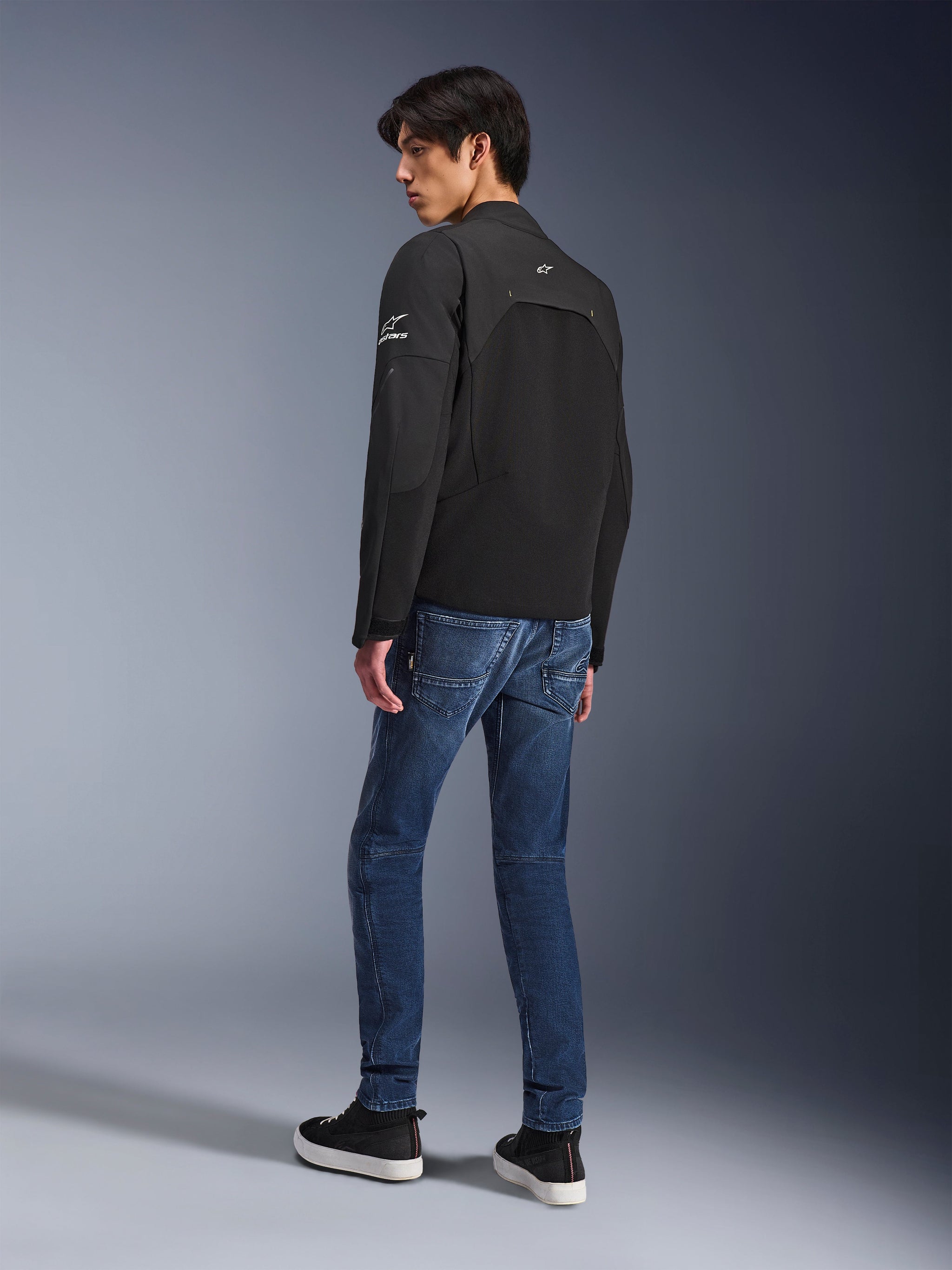 Alpinestars Provoke Jacket, Black urban motorcycle jacket, worn by a model in a rear three-quarter view, paired with blue denim jeans and black sneakers, featuring ripstop softshell inserts and white Alpinestars branding on the sleeve and back.