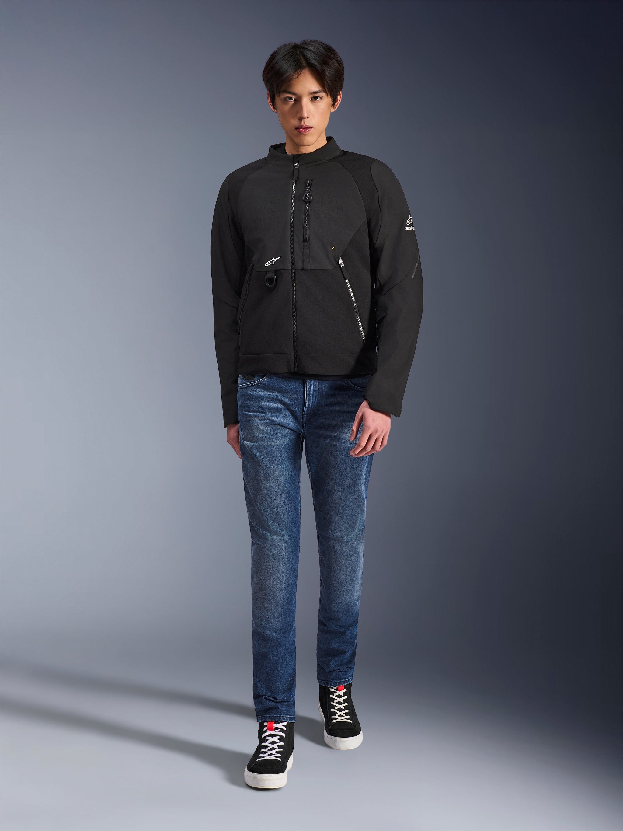 Male model wearing Alpinestars Provoke Jacket in Black, urban textile motorcycle jacket featuring technical zippers and ripstop softshell panels, paired with blue jeans and black sneakers, contemporary urban rider style.