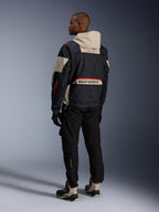 Alpinestars Mogress Air Jacket, Black Almond Hot Orange, back view of rider wearing urban motorcycle gear, almond-colored hood and lower back panel with large Alpinestars branding, orange zip details, paired with black pants and high-top riding boots.