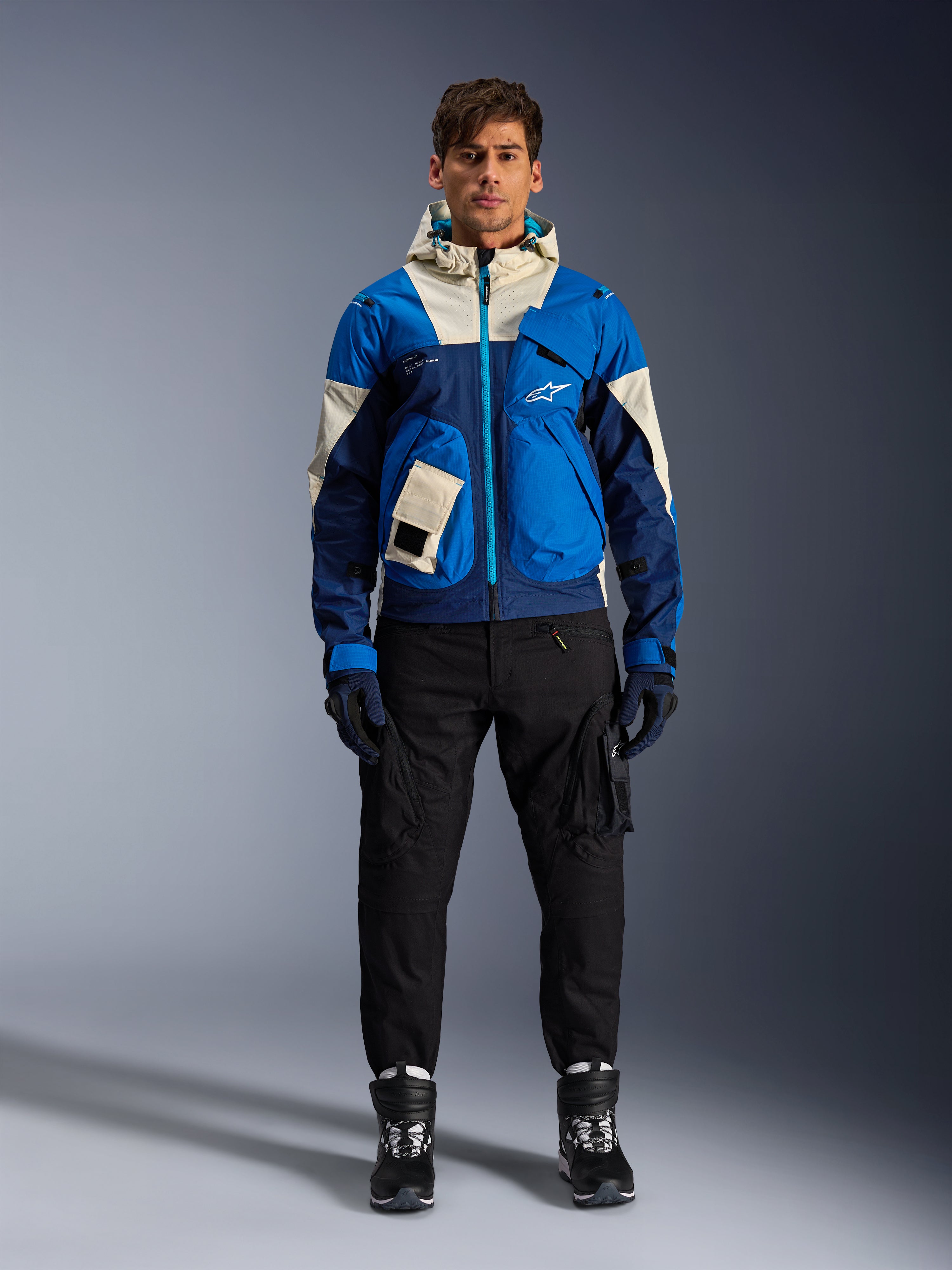 Alpinestars Mogress Air Jacket, Blue Almond urban motorcycle jacket, worn with black pants and boots, featuring a color-blocked blue and cream technical design with utility pockets and a hood, contemporary streetwear-inspired motorcycle apparel