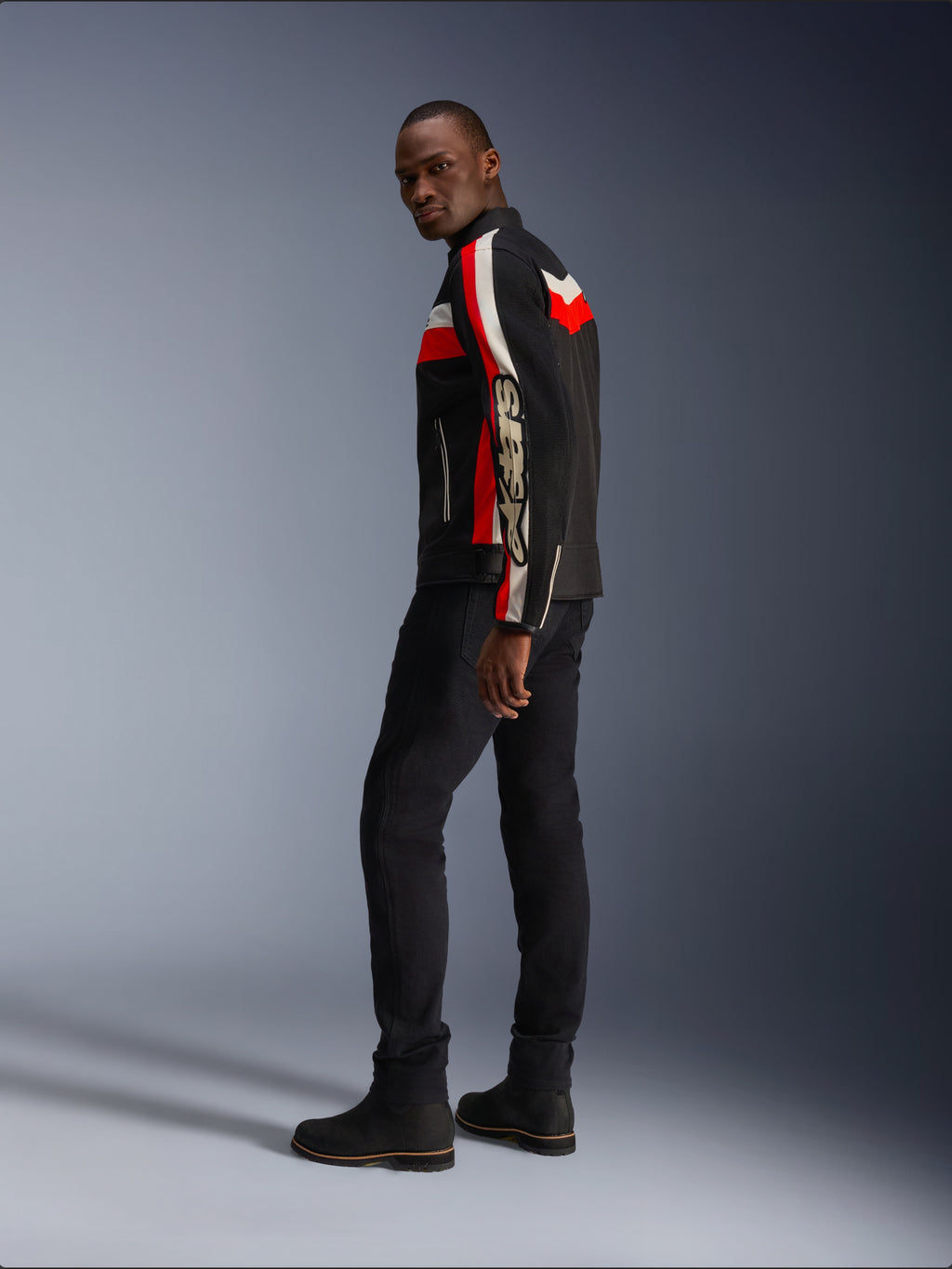 A man wearing the Alpinestars T-Dyno Air Jacket in Black, Bright Red, and Ecru, vintage-inspired textile motorcycle jacket with mesh panels, bold branding on the sleeve, paired with black pants and boots for a modern biker look