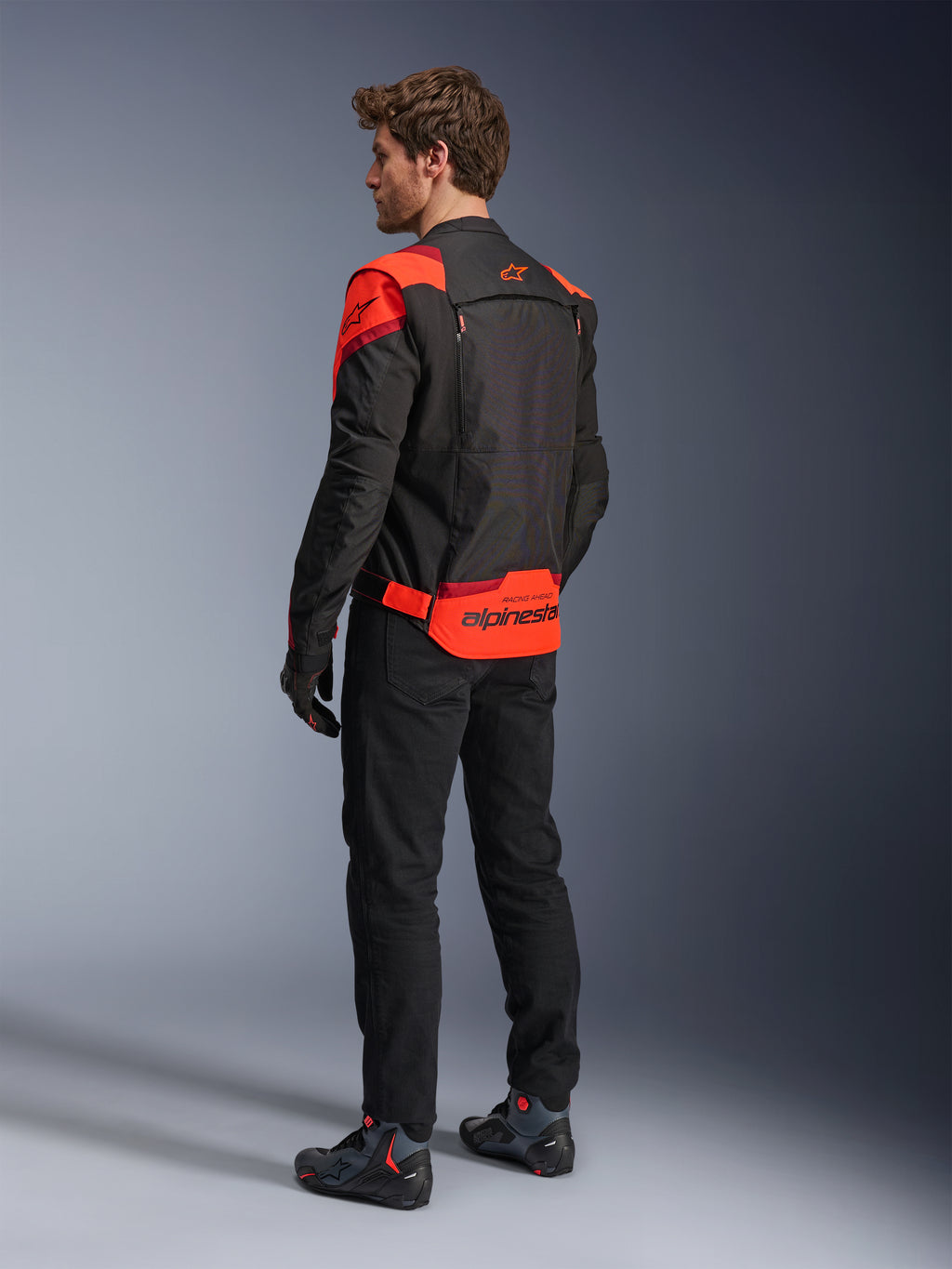 Man wearing Alpinestars T-Stunt Air Jacket from the back, Black Red Fluo and Dark Red, textile motorcycle jacket with mesh panels and sport-cut design, featuring bold Alpinestars branding on the lower back, styled with black pants and riding boots