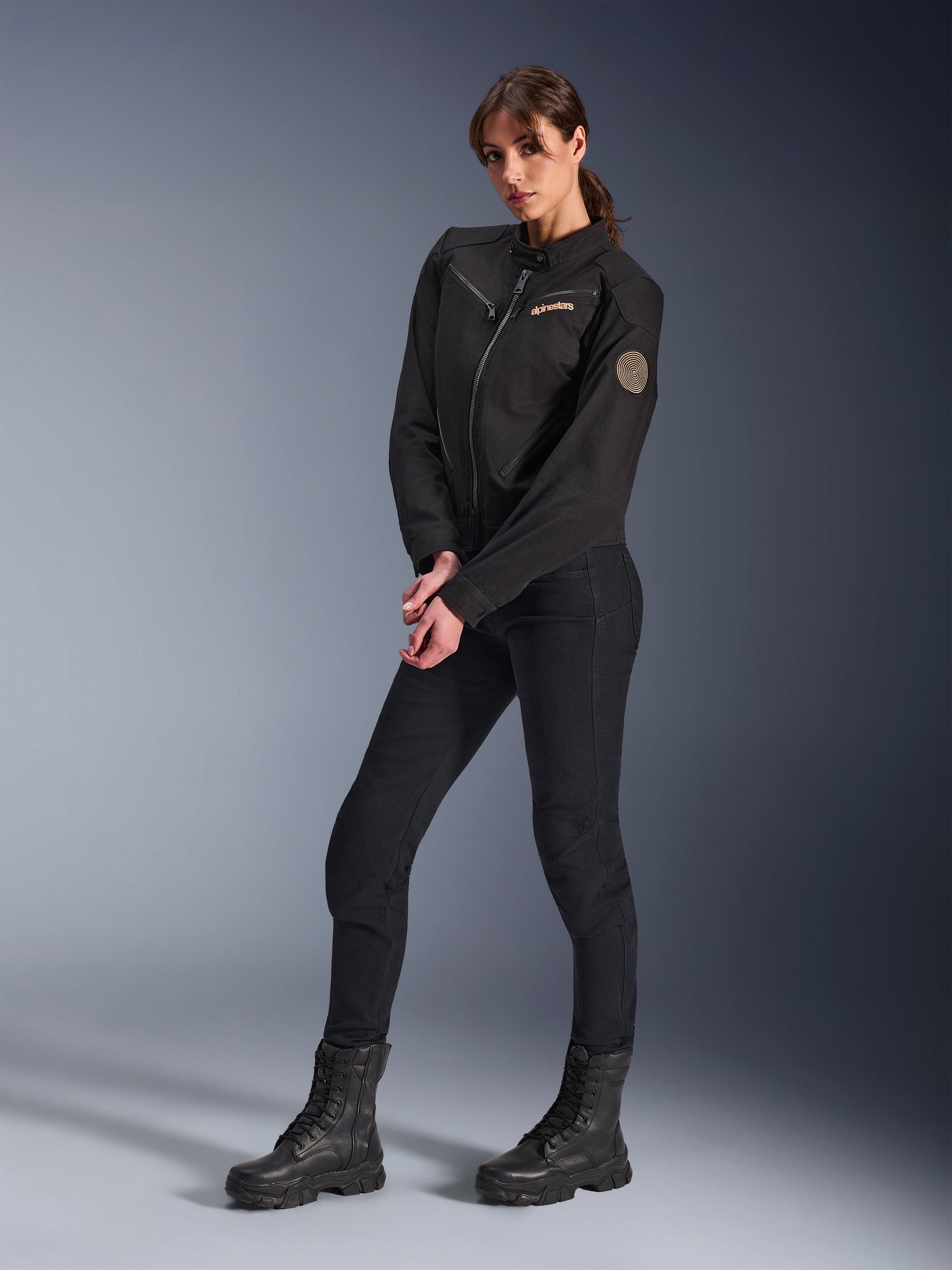 Female model wearing the Alpinestars Woman Stella Grace Jacket, Black Urban Motorcycle Jacket, styled with black riding pants and boots, workwear-inspired design featuring a bronze embroidered logo on the chest and a circular spiral patch on the sleeve