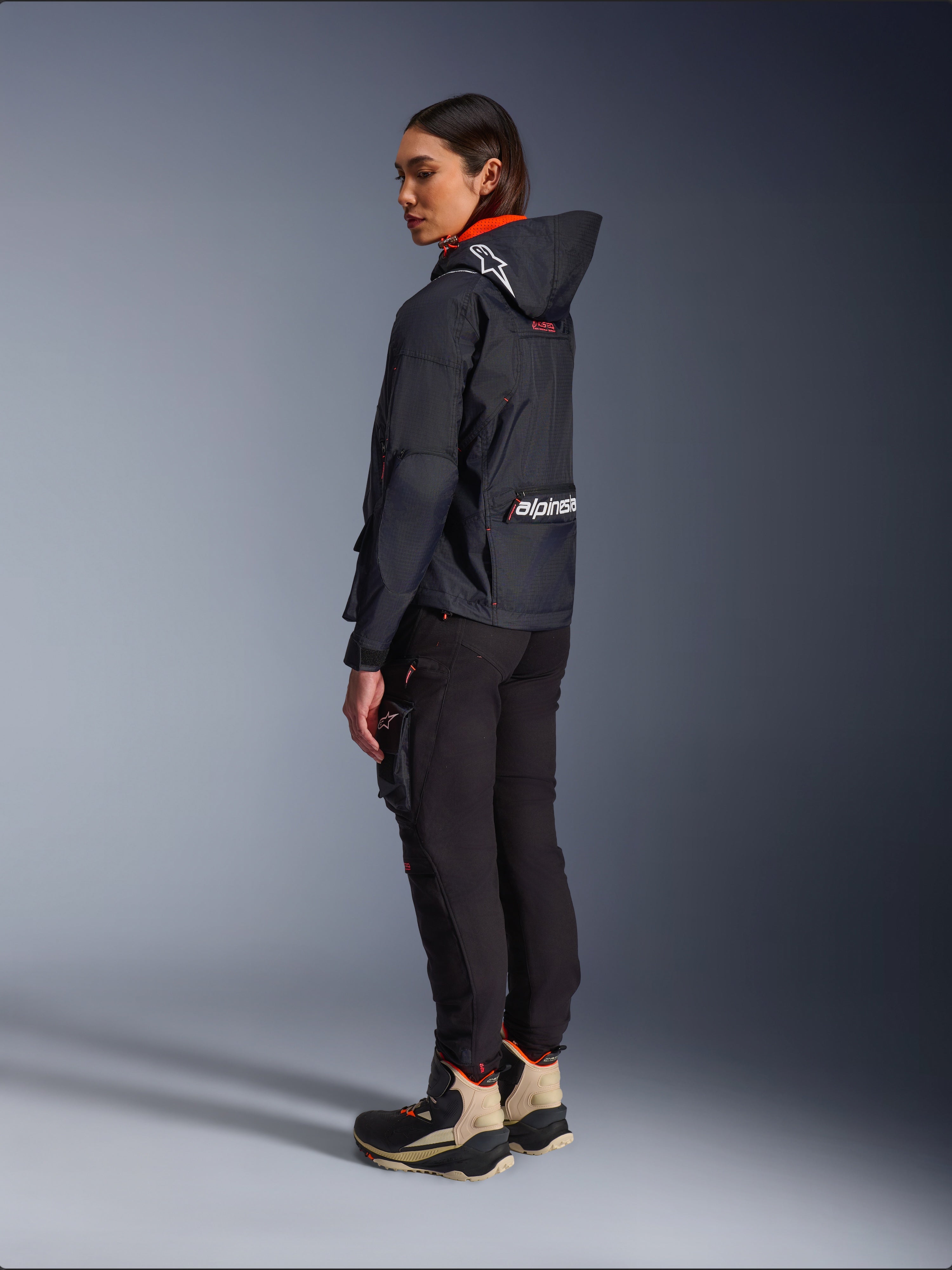 Female rider wearing the Alpinestars Molly Air Jacket in Black Fluo Coral, rear side view showing the hood with Astars logo and large Alpinestars branding on the back, paired with black cargo riding pants and tan and black adventure boots, contemporary urban motorcycle gear for women
