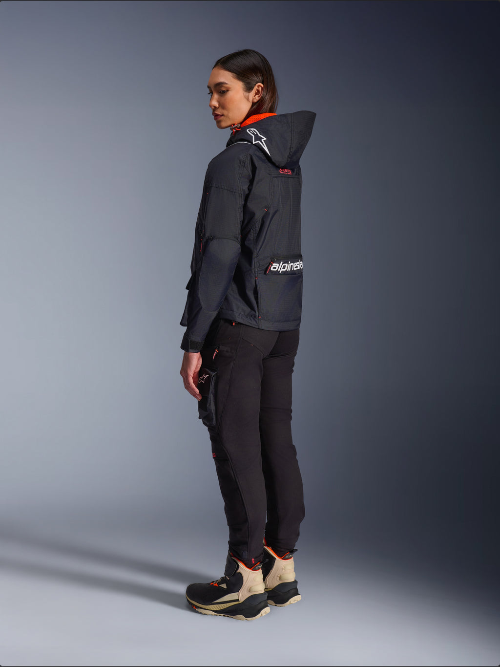 Alpinestars Actea Women's Pants, Black, modern motorcycle pants with a tapered fit and thigh cargo pocket, shown from a side-rear perspective on a model wearing a dark hooded jacket and riding boots, urban riding style.