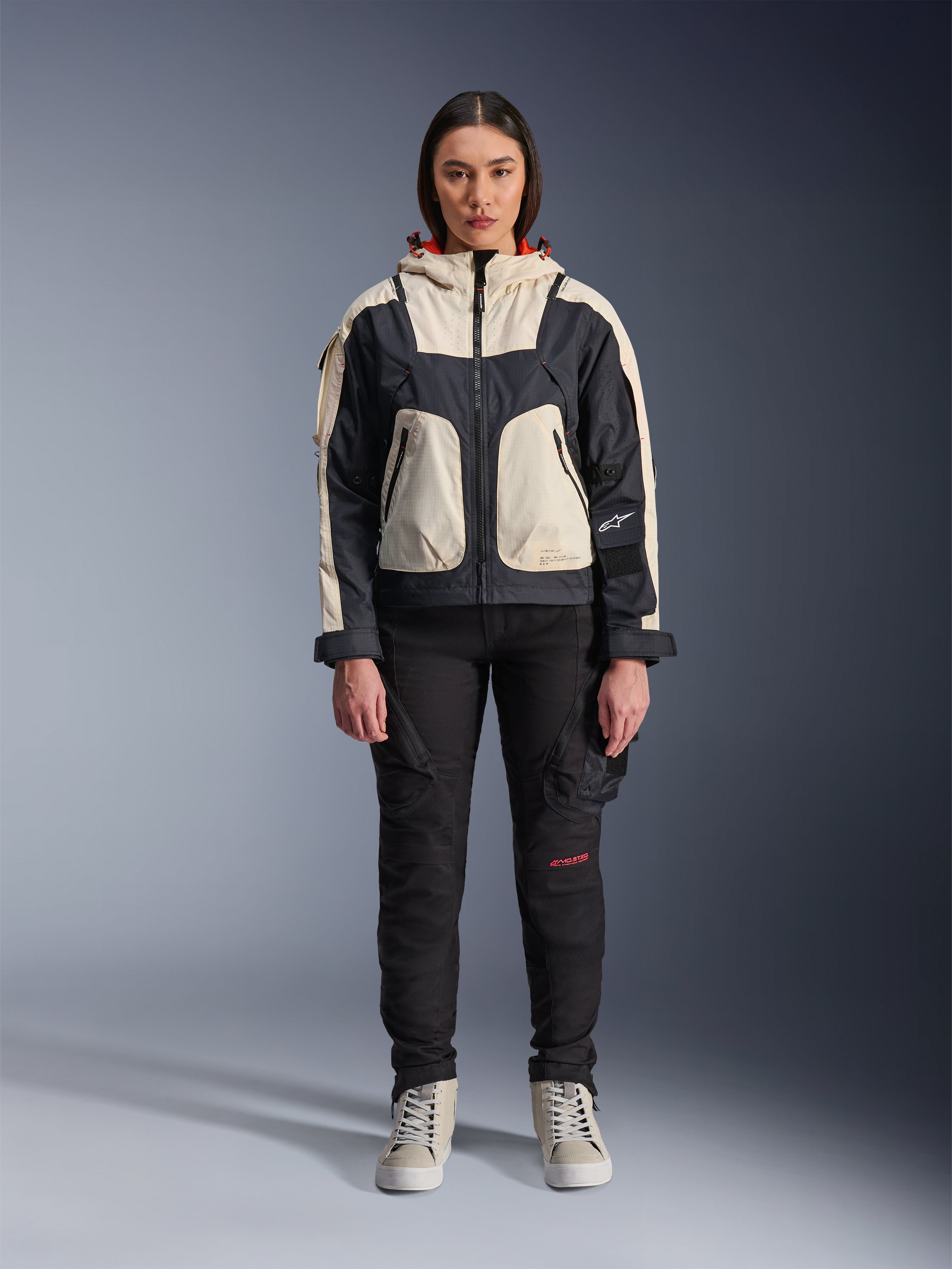Female model wearing the Alpinestars Molly Air Jacket in Black Almond, featuring a color-blocked design with cream and black panels and a red-lined hood, paired with black motorcycle pants and cream sneakers, studio shot of urban contemporary riding gear.