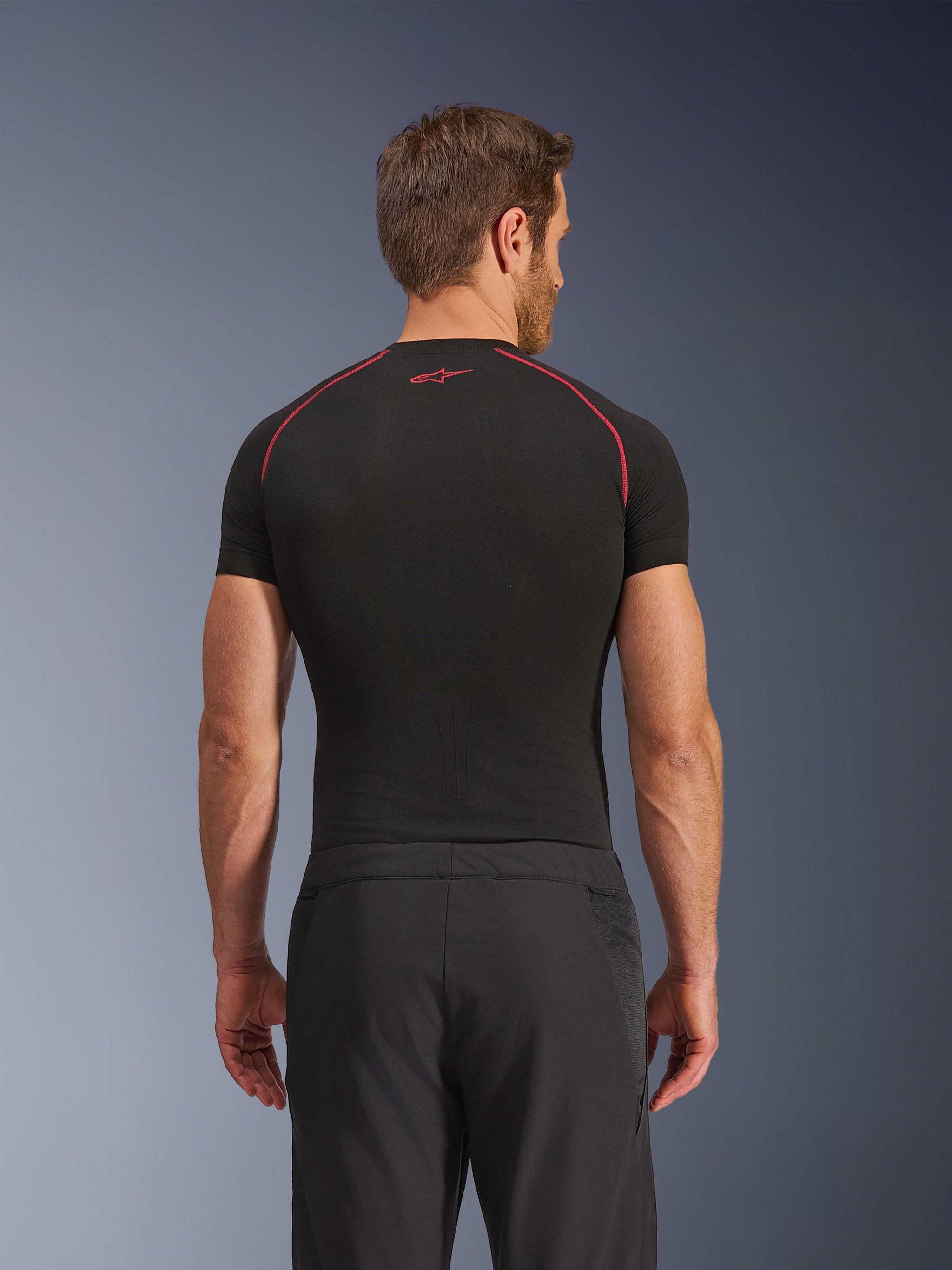 Rear view of a model wearing Alpinestars Aeroshell Airflow Pants, Black, lightweight softshell construction with integrated mesh side panels, paired with a black base layer featuring red contrast stitching, sporty motorcycle attire