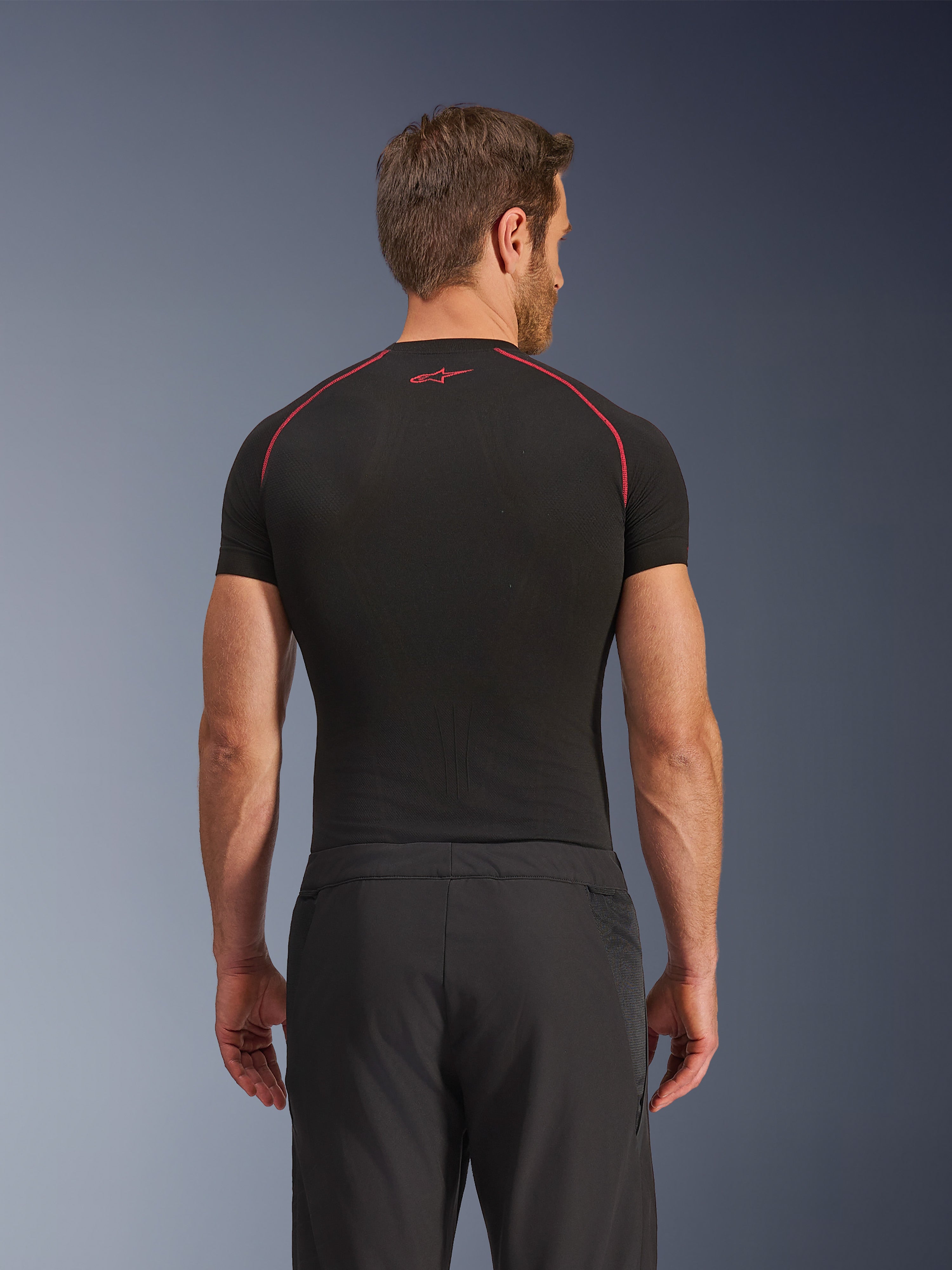 Rear view of a model wearing Alpinestars Aeroshell Airflow Pants, Black, lightweight softshell construction with integrated mesh side panels, paired with a black base layer featuring red contrast stitching, sporty motorcycle attire