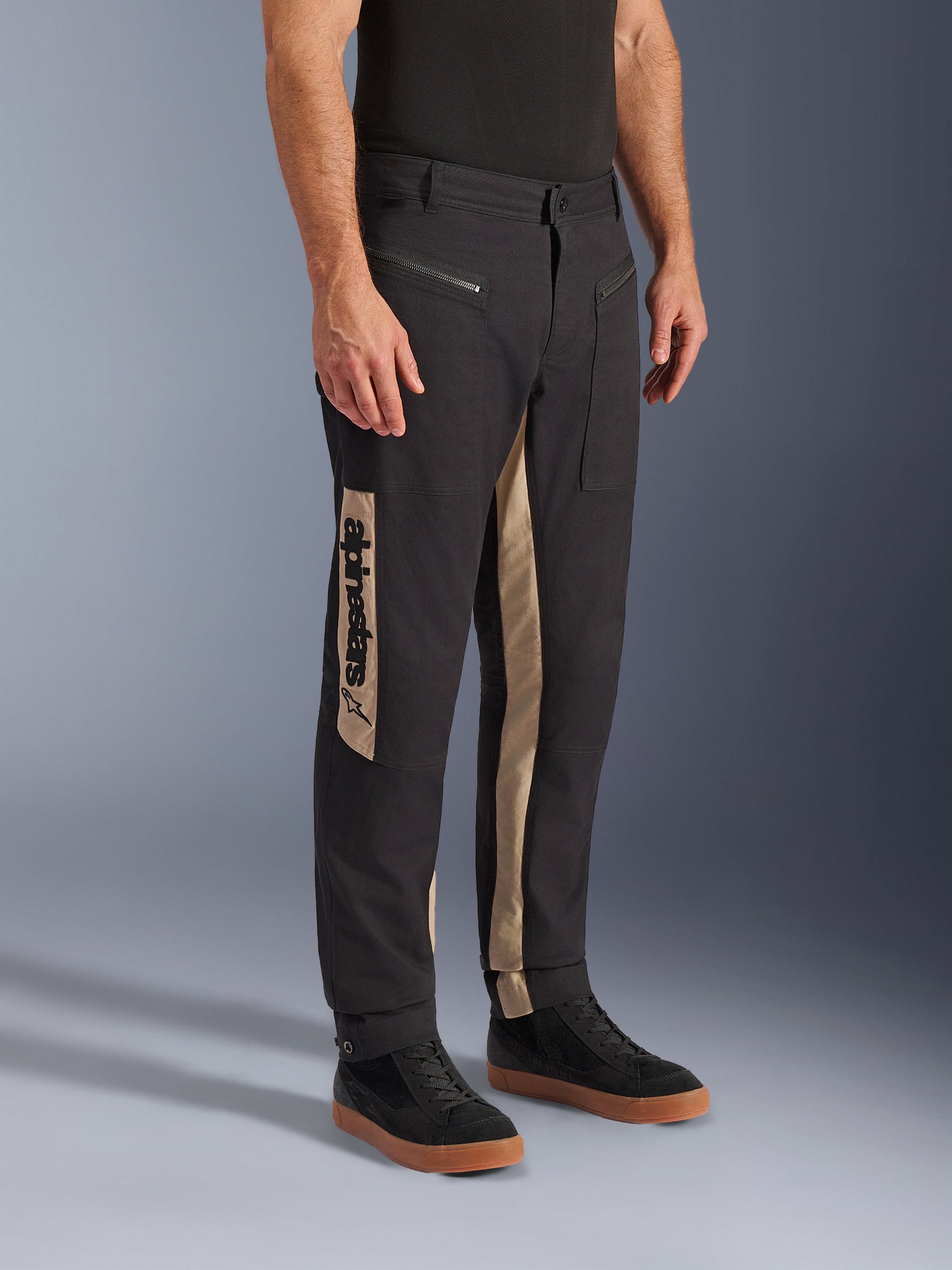 Alpinestars Flex-AST Canvas Pants, Urban Motorcycle Pants, Black Aluminum, featuring tan side panels with a vertical Alpinestars logo, workwear-inspired design with zippered pockets and ergonomic cut, worn with casual black sneakers for a modern city look.