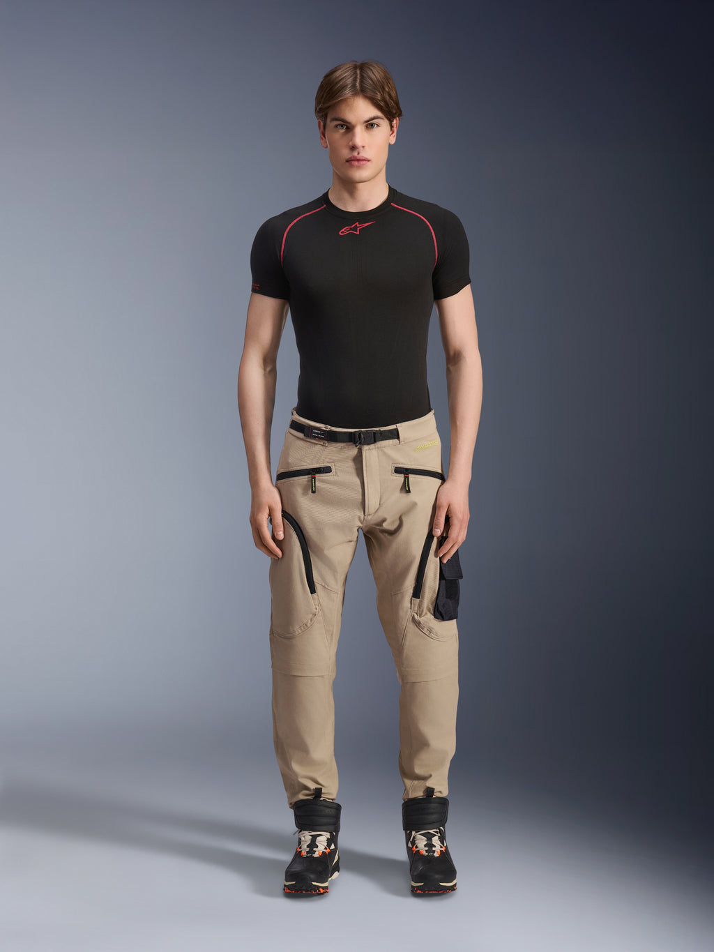 Male model wearing Alpinestars Acteon Pants in Sand, paired with a black short-sleeve base layer and black technical boots, featuring cargo pockets with black zippers and a tapered fit, contemporary urban motorcycle gear
