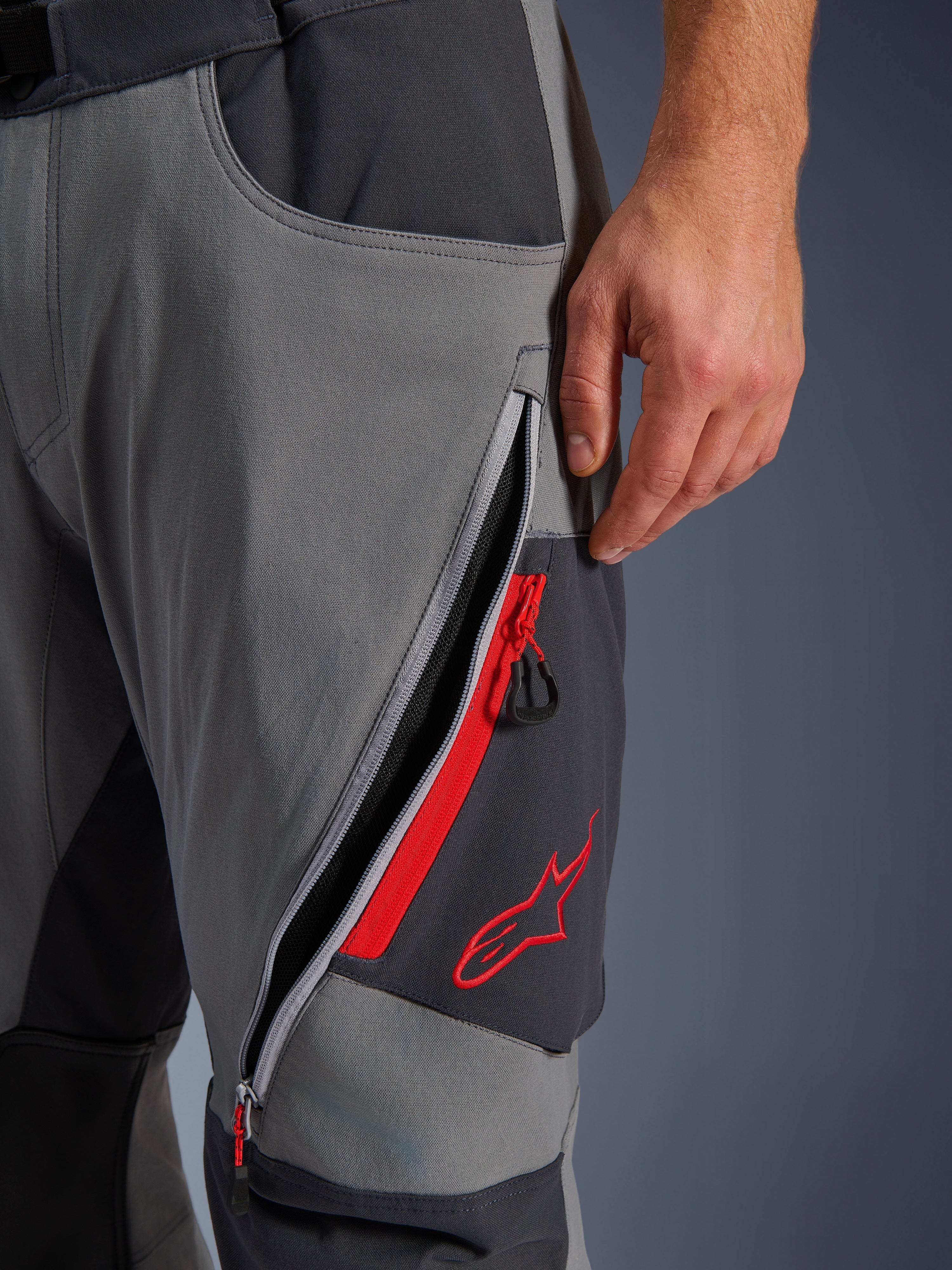 Alpinestars Maxdura Dual Pants, Dark Gray and Deep Gray adventure motorcycle pants, close-up view of open zippered thigh vent with red highlights and embroidered red star logo, technical stretch fabric design