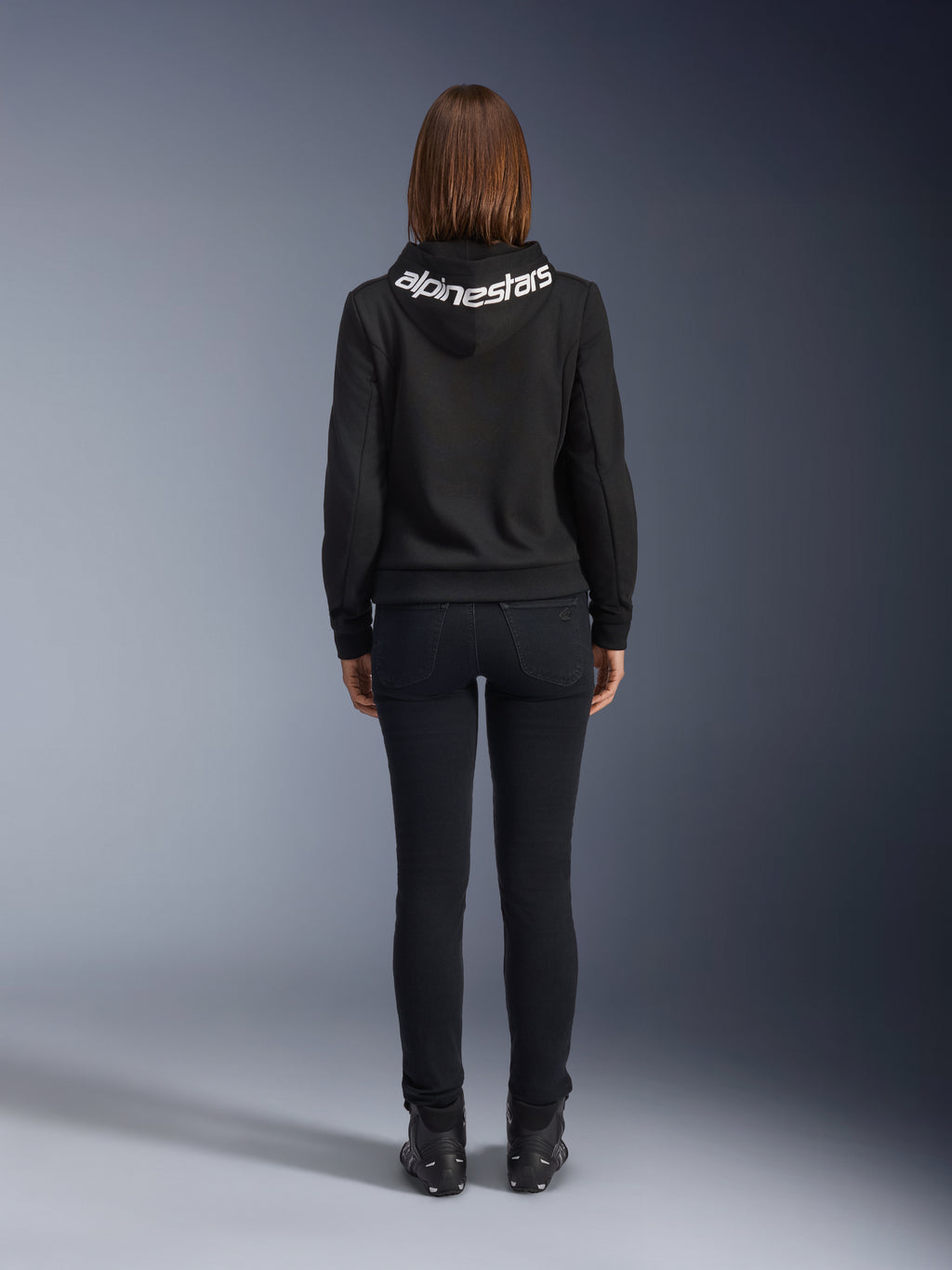 Daisy V3 Women's Riding Denim, Black Rinse, slim fit motorcycle pants, rear view showing high back waist and feminine silhouette, paired with a black Alpinestars hoodie and black riding boots, urban casual riding gear