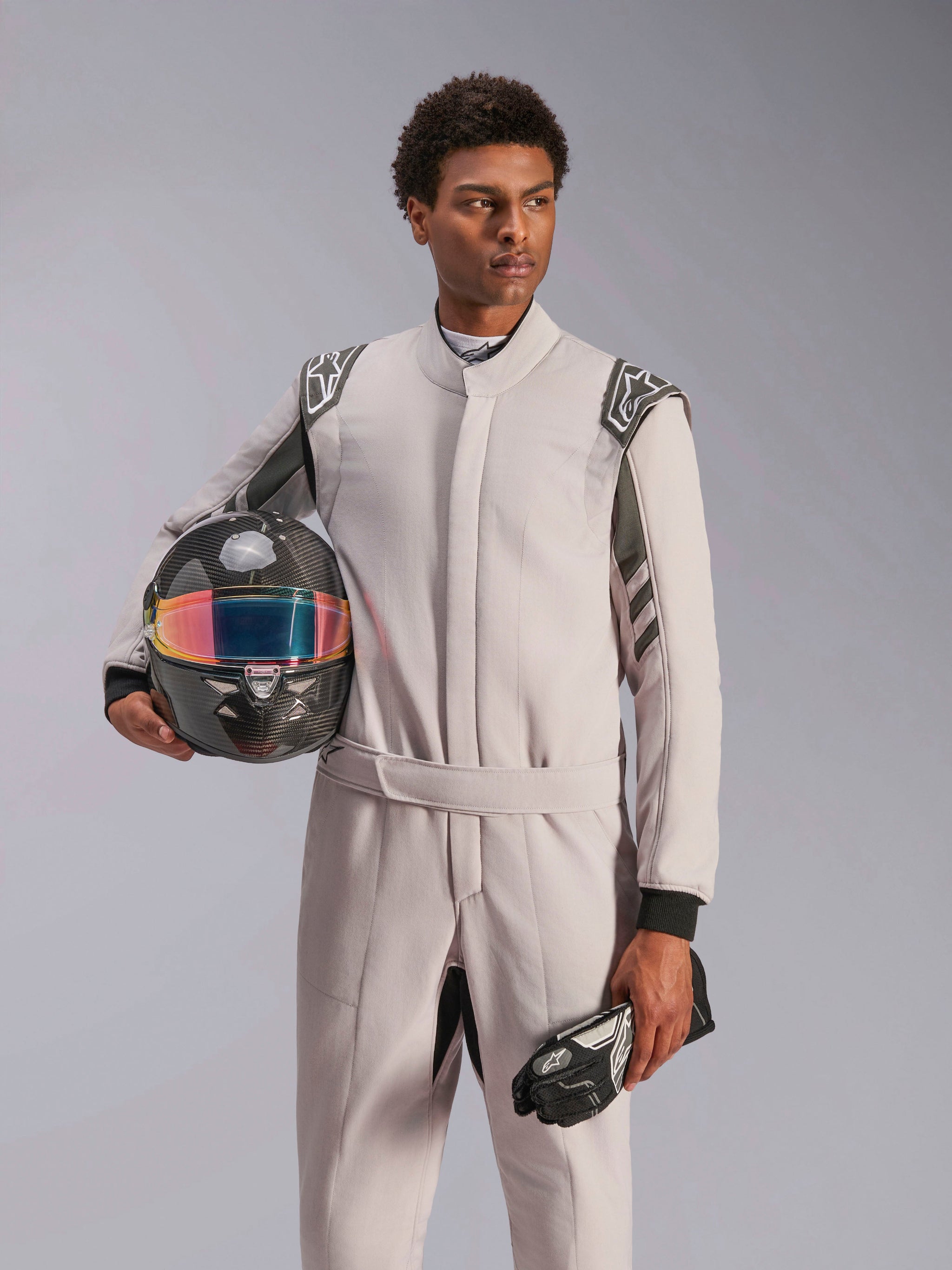 Alpinestars GP Race V3 Suit FIA, Auto Racing Suit, Silver Anthracite, worn by model holding a carbon fiber helmet with an iridescent visor and black racing gloves, professional motorsport protective gear with visible shoulder epaulettes.