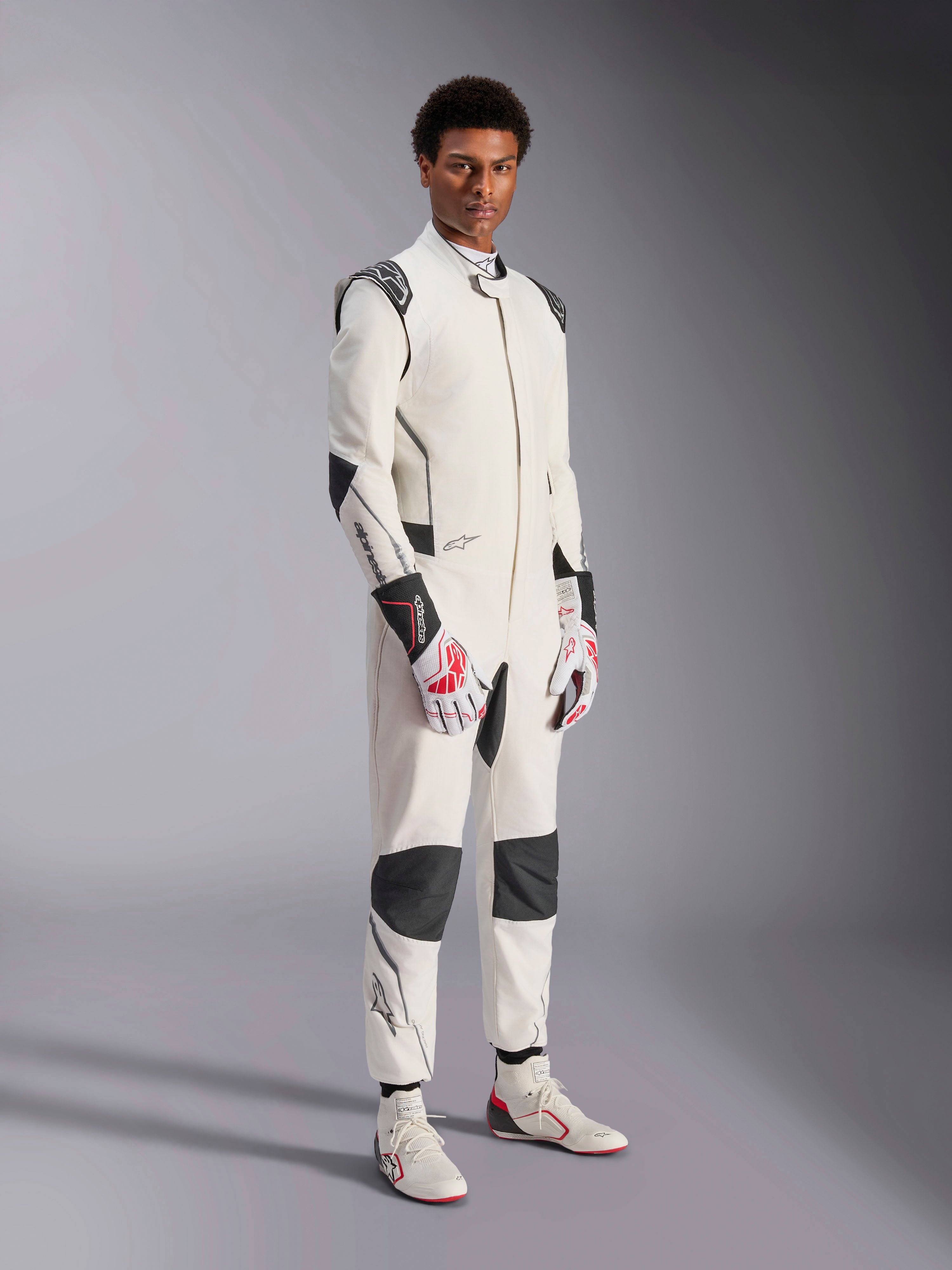 Male model wearing the Alpinestars Hypertech V3 Suit, White with Black and Silver accents, professional auto racing gear featuring black panels on shoulders and knees, styled with white and red racing gloves and white racing boots