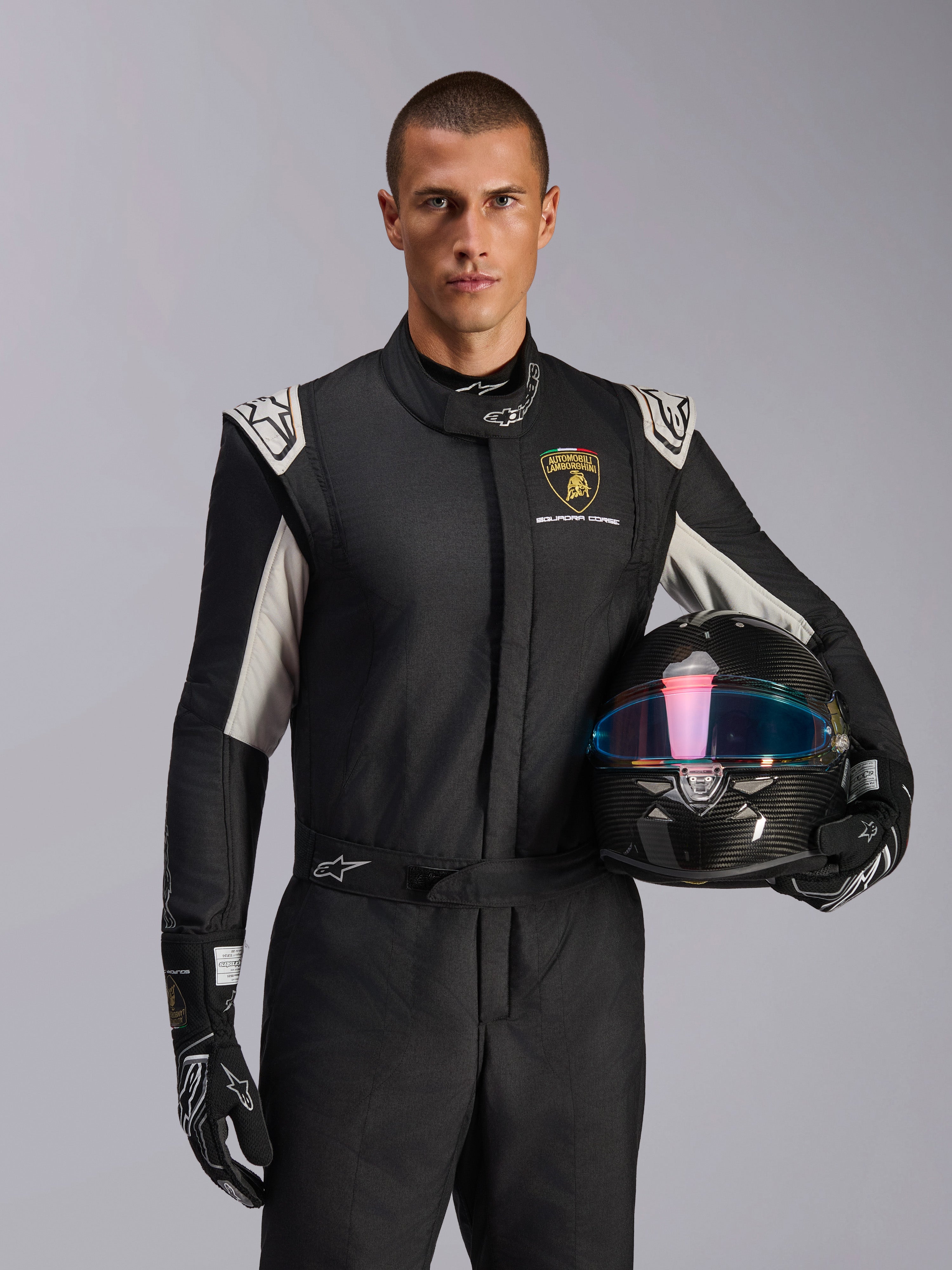 Alpinestars Limited Edition Lamborghini GP Tech V4 Suit, Auto Racing Suit, Black Silver and Gold, worn by a model holding a carbon fiber helmet with an iridescent visor, featuring Lamborghini Squadra Corse branding and white shoulder accents