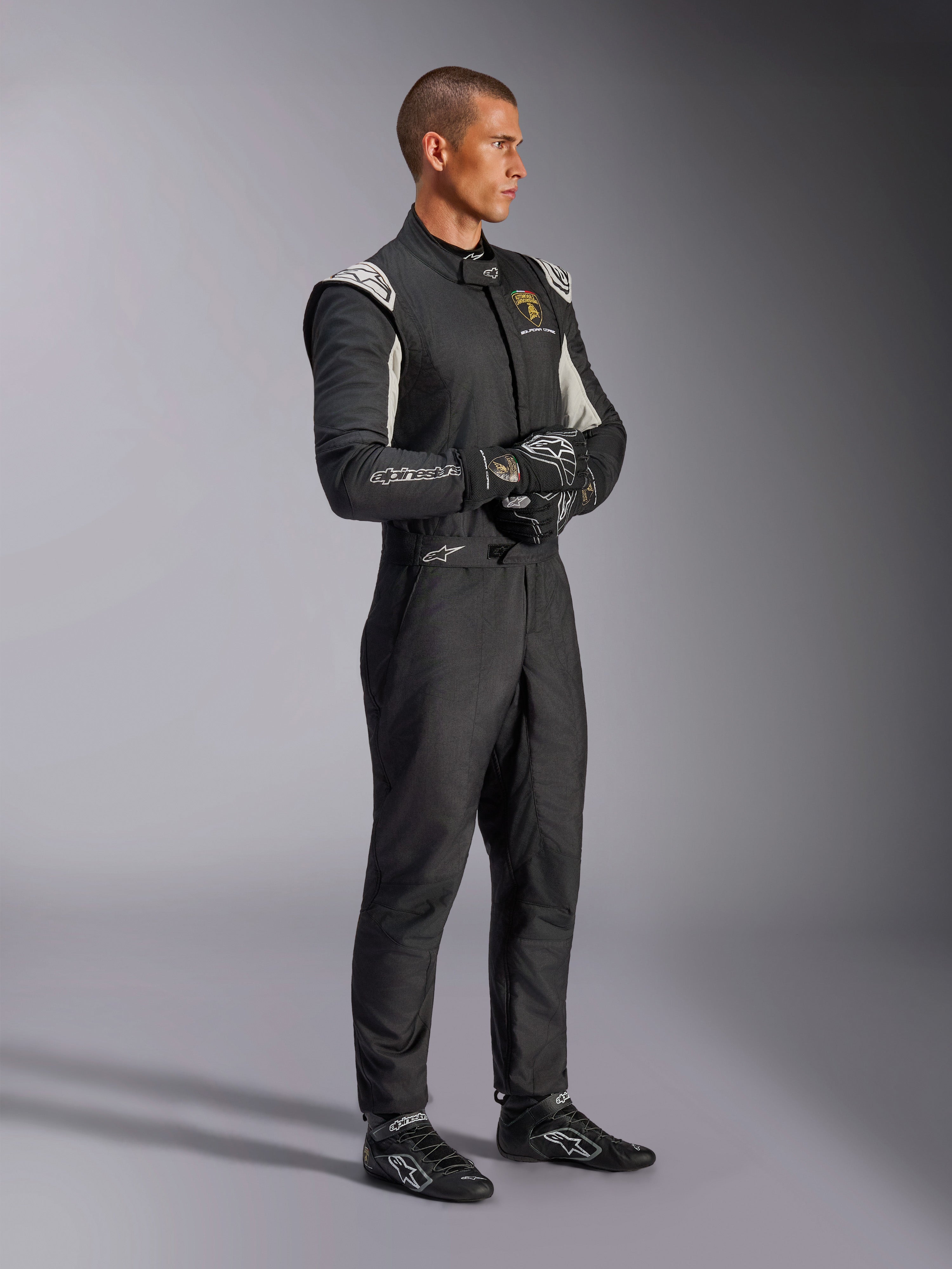 Male driver wearing the Alpinestars Limited Edition Lamborghini GP Tech V4 Suit, FIA-certified auto racing suit in Black, Silver, and Gold, paired with matching racing gloves and shoes, featuring Lamborghini Squadra Corse branding, professional motorsport racing gear